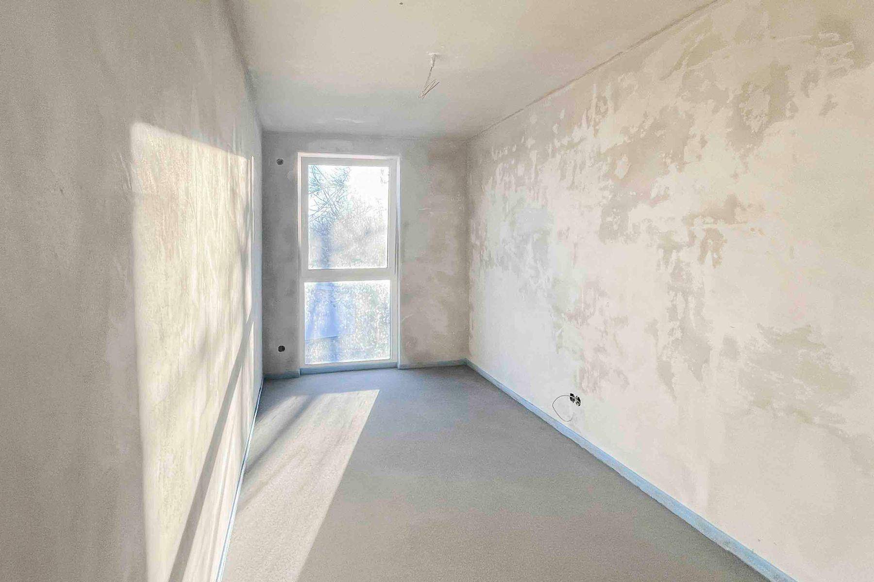 Heller Rohbau-Raum mit Estrichboden und Sonneneinstrahlung durch das Fenster.