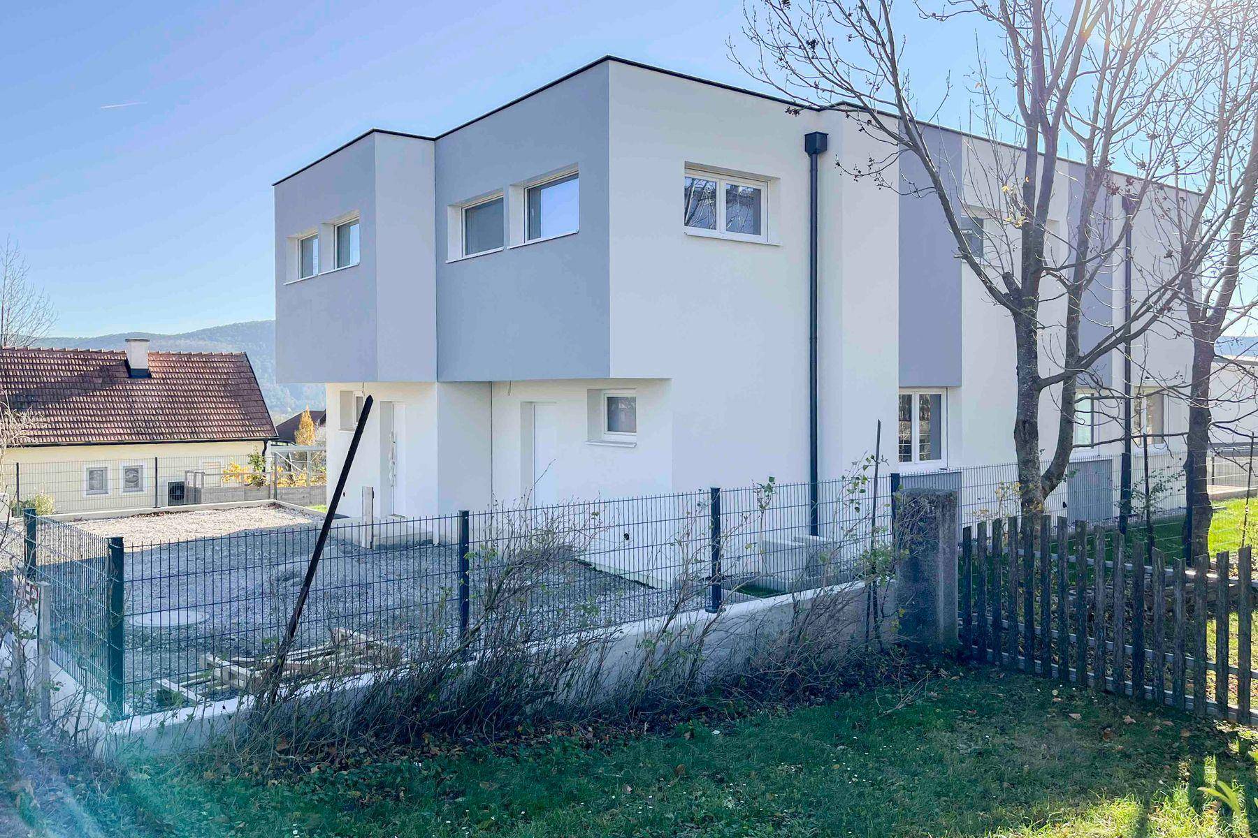 Seitenansicht des modernen Neubaus mit umzäuntem Grundstück und Blick auf die Umgebung.