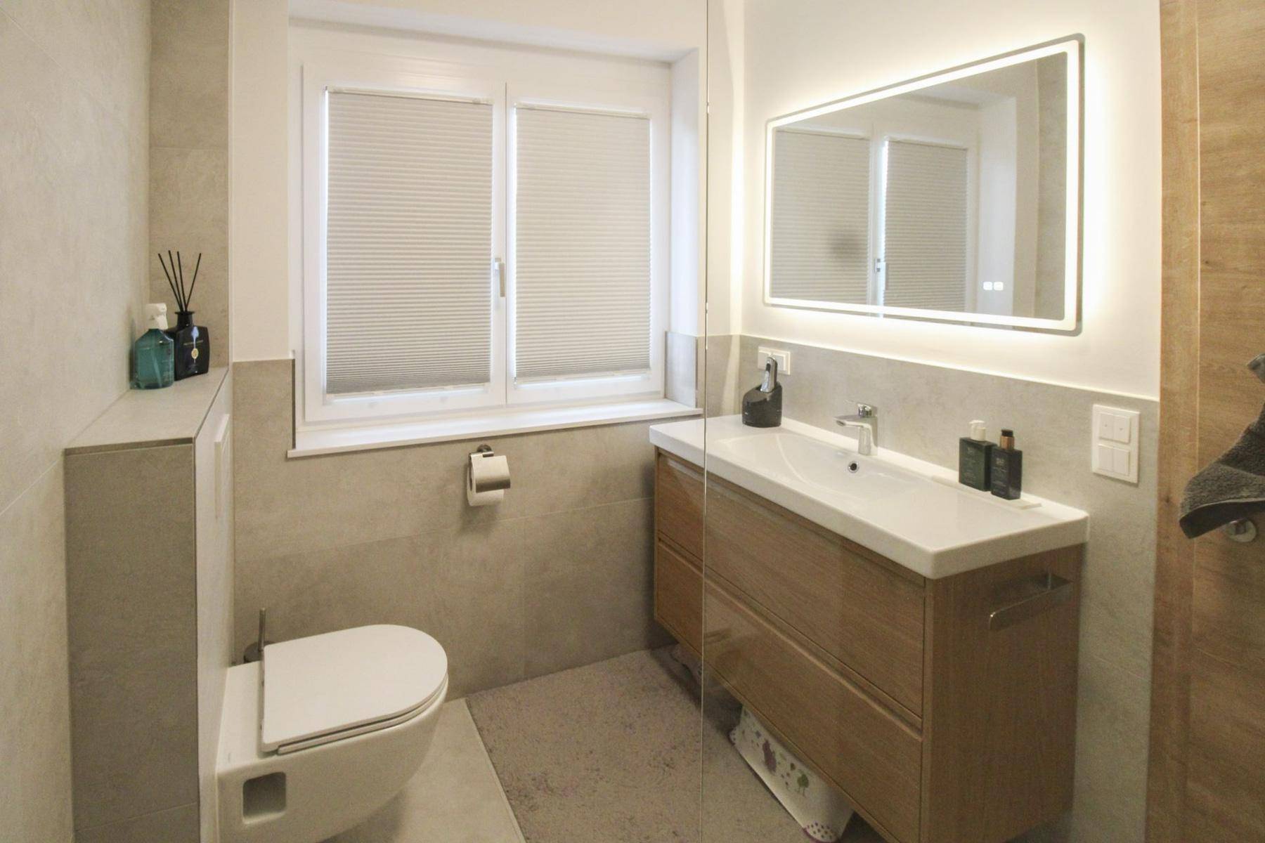Modernes Badezimmer mit wandhängendem WC, Einzelwaschbecken und beleuchtetem Spiegel.