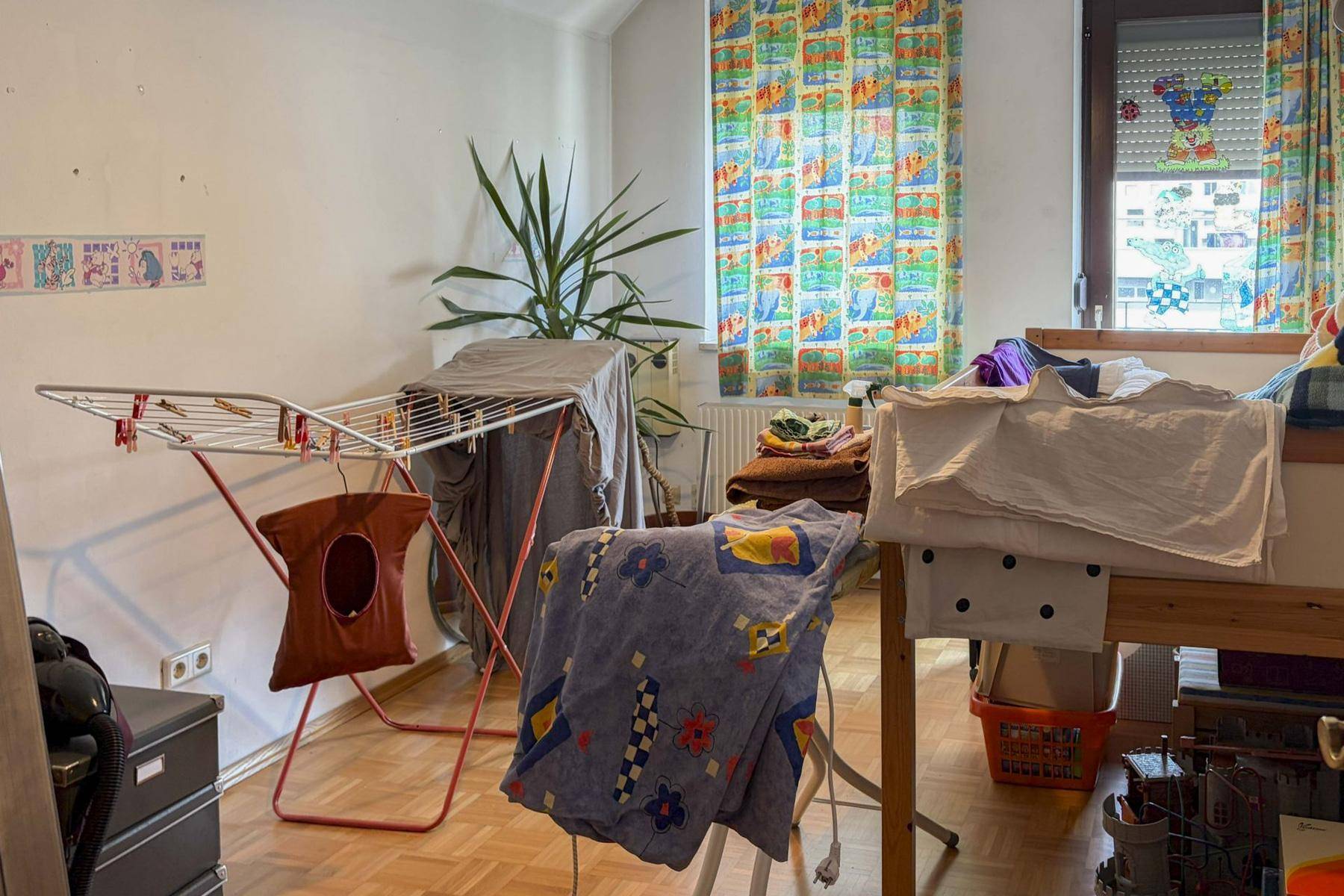 Zimmer mit Wäscheständer, Pflanzen und bunten Vorhängen, möglicherweise als Hauswirtschaftsraum genutzt.