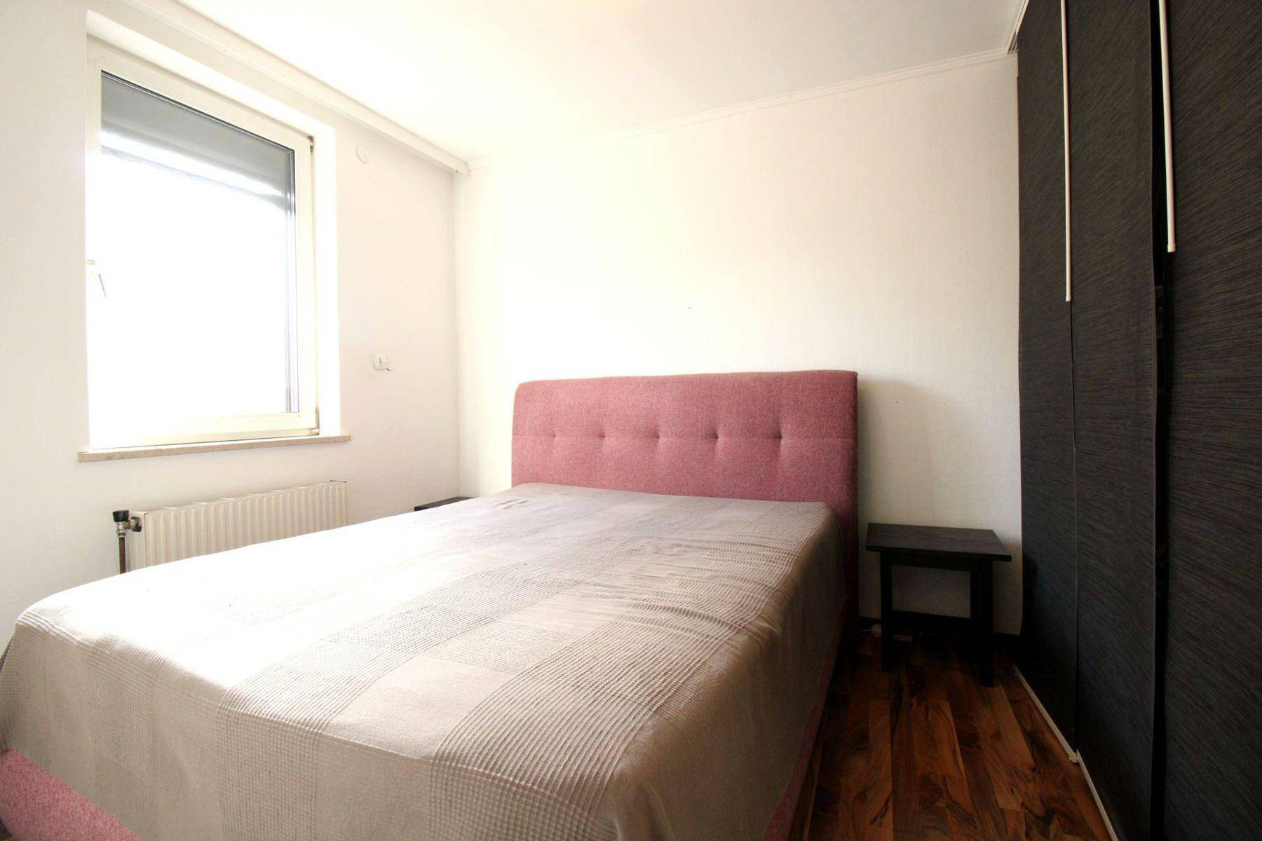 Schlafzimmer mit rosa gepolstertem Bett, dunklem Kleiderschrank und Fenster mit Rollladen.
