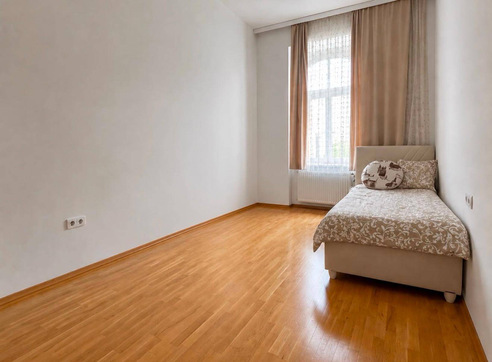 Einfaches Zimmer mit Einzelbett, Holzboden und Fenster mit beigen Vorhängen.