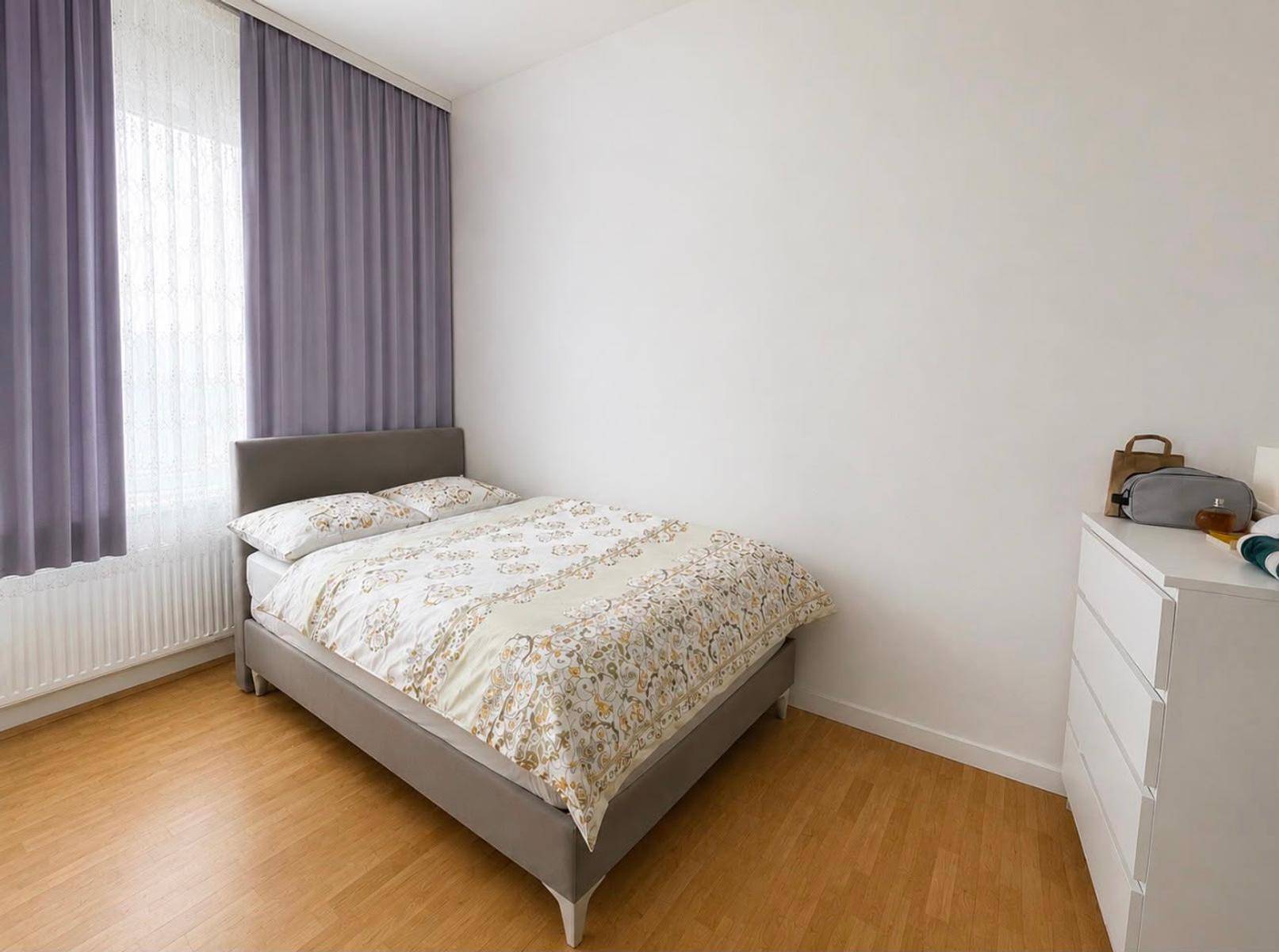 Schlichtes Schlafzimmer mit Doppelbett, Holzboden und großem Fenster mit lila Vorhängen.
