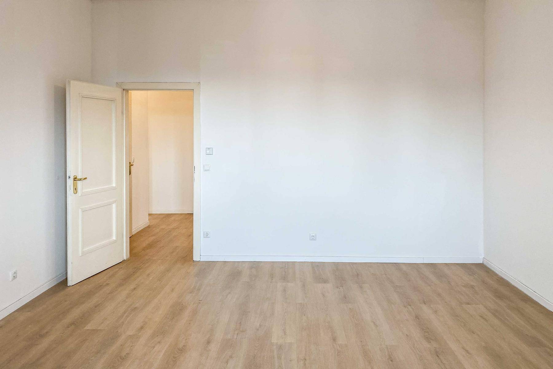 Helles, leeres Zimmer mit Laminatboden und einer Tür, die zu einem weiteren Raum führt.