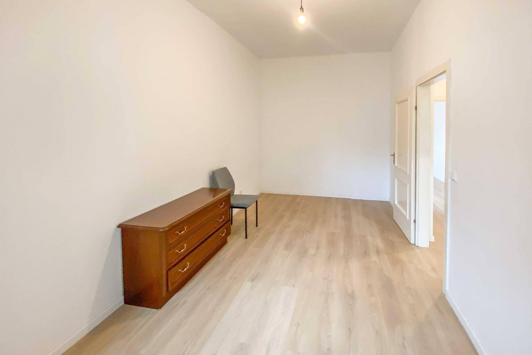 Geräumiges, helles Zimmer mit Laminatboden, weißen Wänden und einfacher Möblierung.