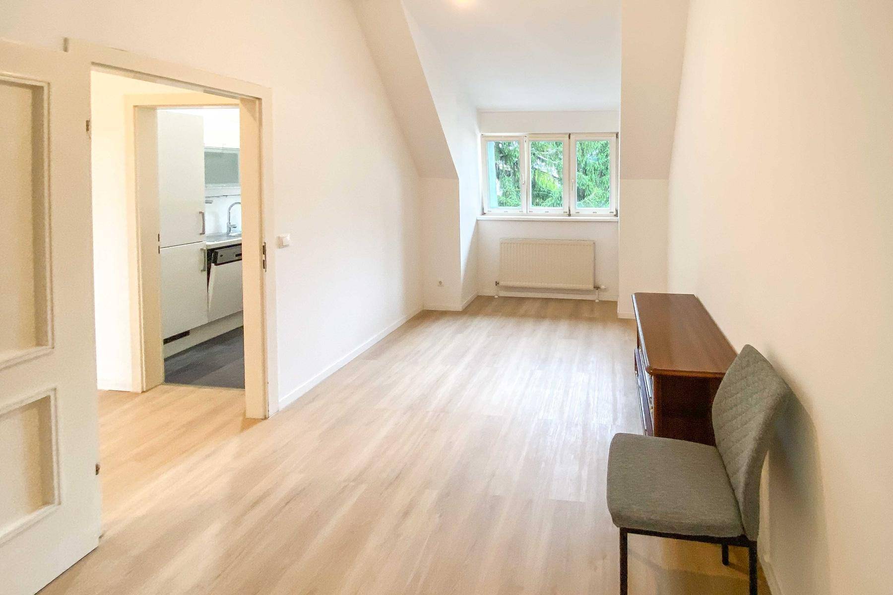 Heller Raum mit Dachschräge und Fenster, angrenzend an eine Küche, mit einfacher Einrichtung.