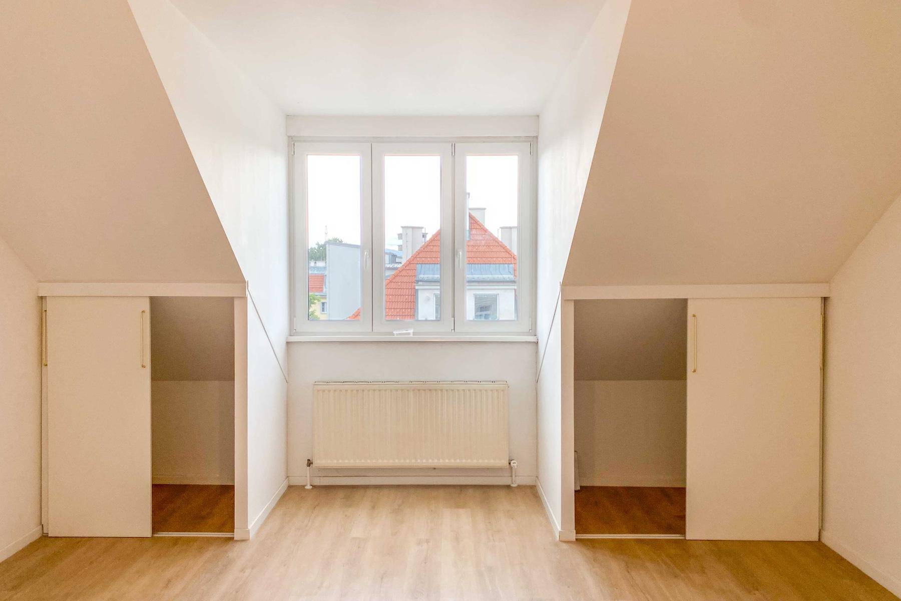 Fensterbereich in einem Zimmer mit Dachschrägen, Einbauschränken und Blick auf Dächer.