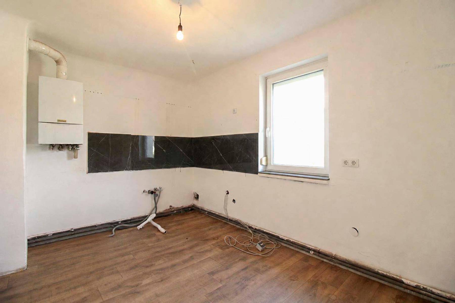 Leere Küche mit Fenster, Gastherme und vorbereiteten Anschlüssen für Einbaumöbel.