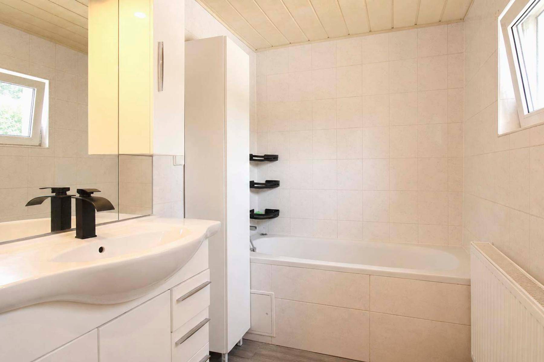 Badezimmer mit Badewanne, Waschtisch, Spiegelschrank und zwei Fenstern.