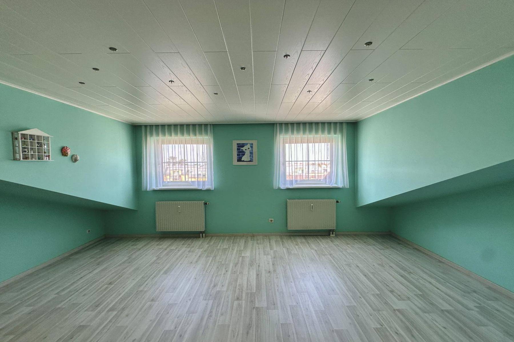 Helles Zimmer mit mintgrünen Wänden, Laminatboden und zwei Fenstern, die viel Licht spenden.