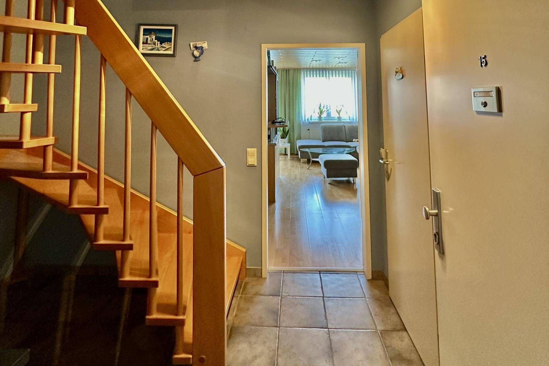 Eingangsbereich mit Fliesenboden, Holztreppe und Durchgang zu einem hellen Wohnzimmer.