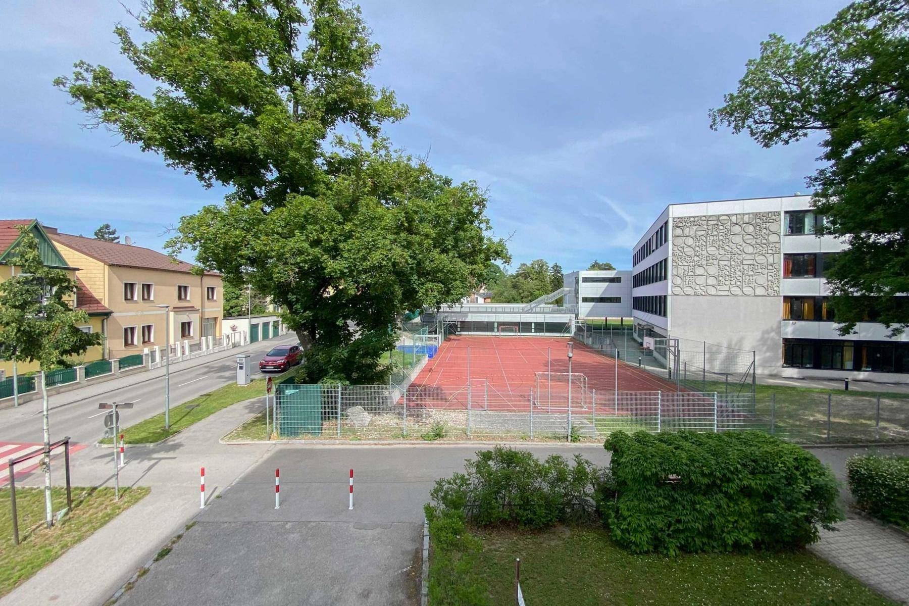 Weitläufiger Blick auf einen Sportplatz, umliegende Gebäude und grüne Bäume unter blauem Himmel.
