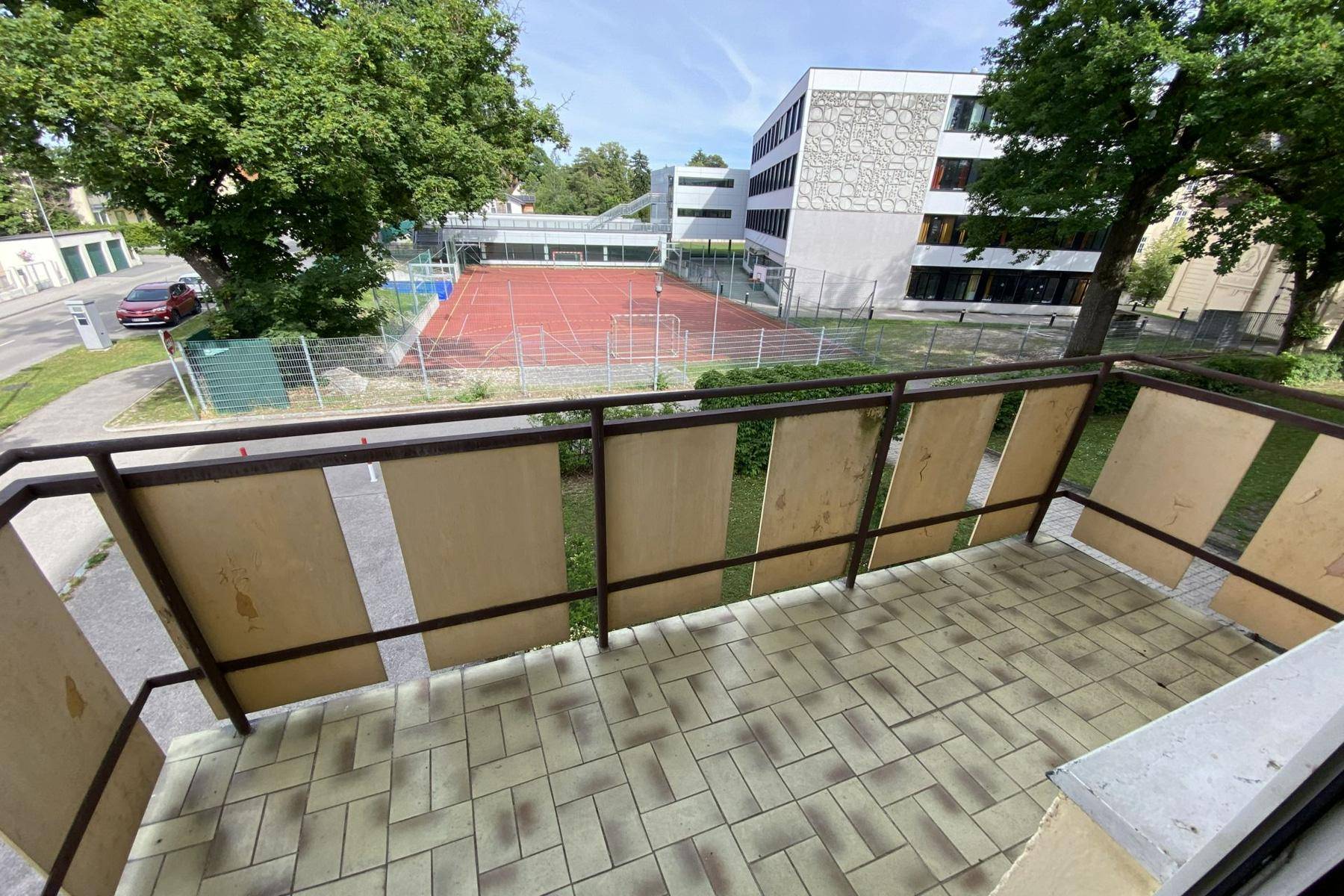 Balkon mit Fliesenboden und Blick auf einen Sportplatz und umliegende Gebäude.