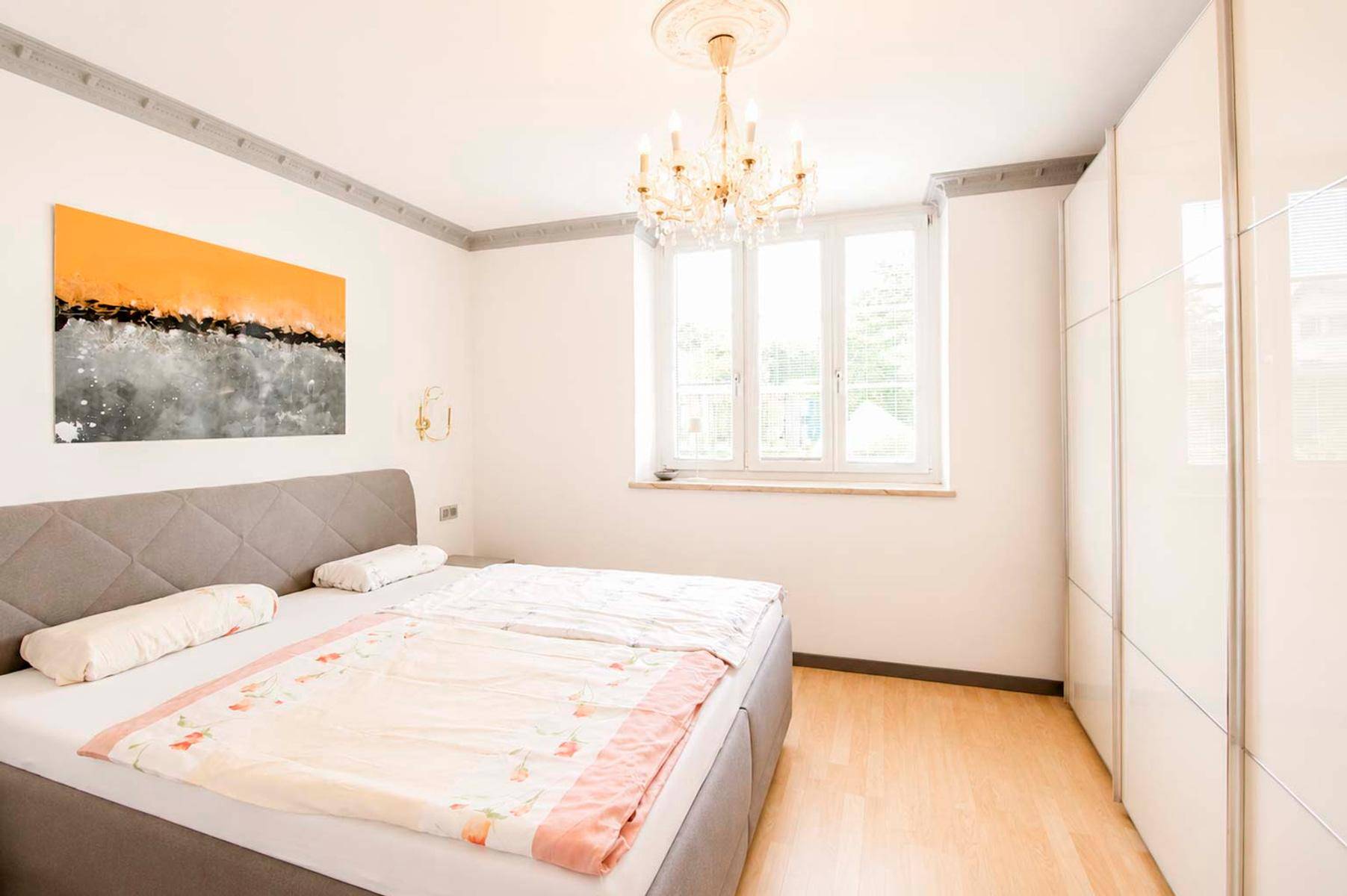 Helles Schlafzimmer mit Doppelbett, Kronleuchter, Einbauschrank und Stuckdecke.