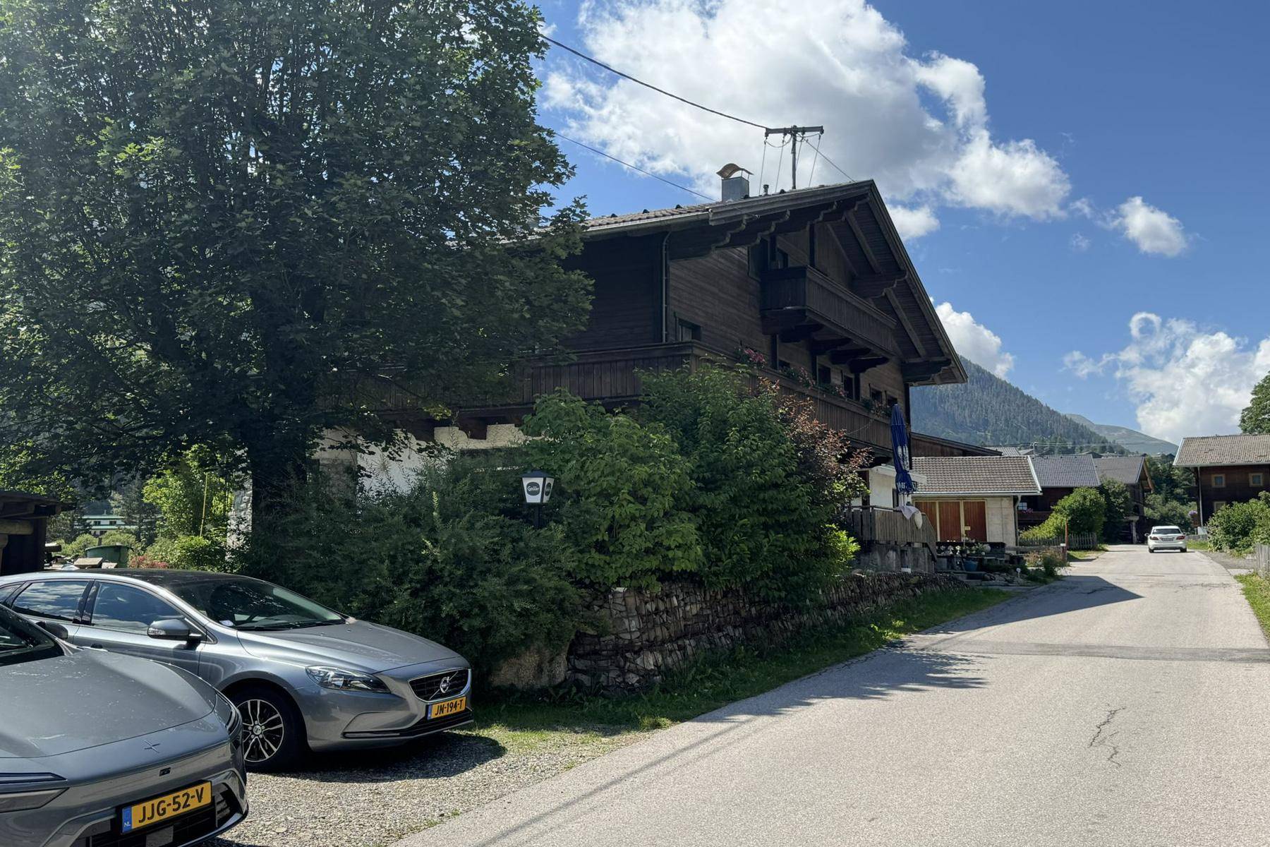 Seitenansicht des traditionellen Holzhauses mit geparkten Autos und einer Bergkulisse im Hintergrund.