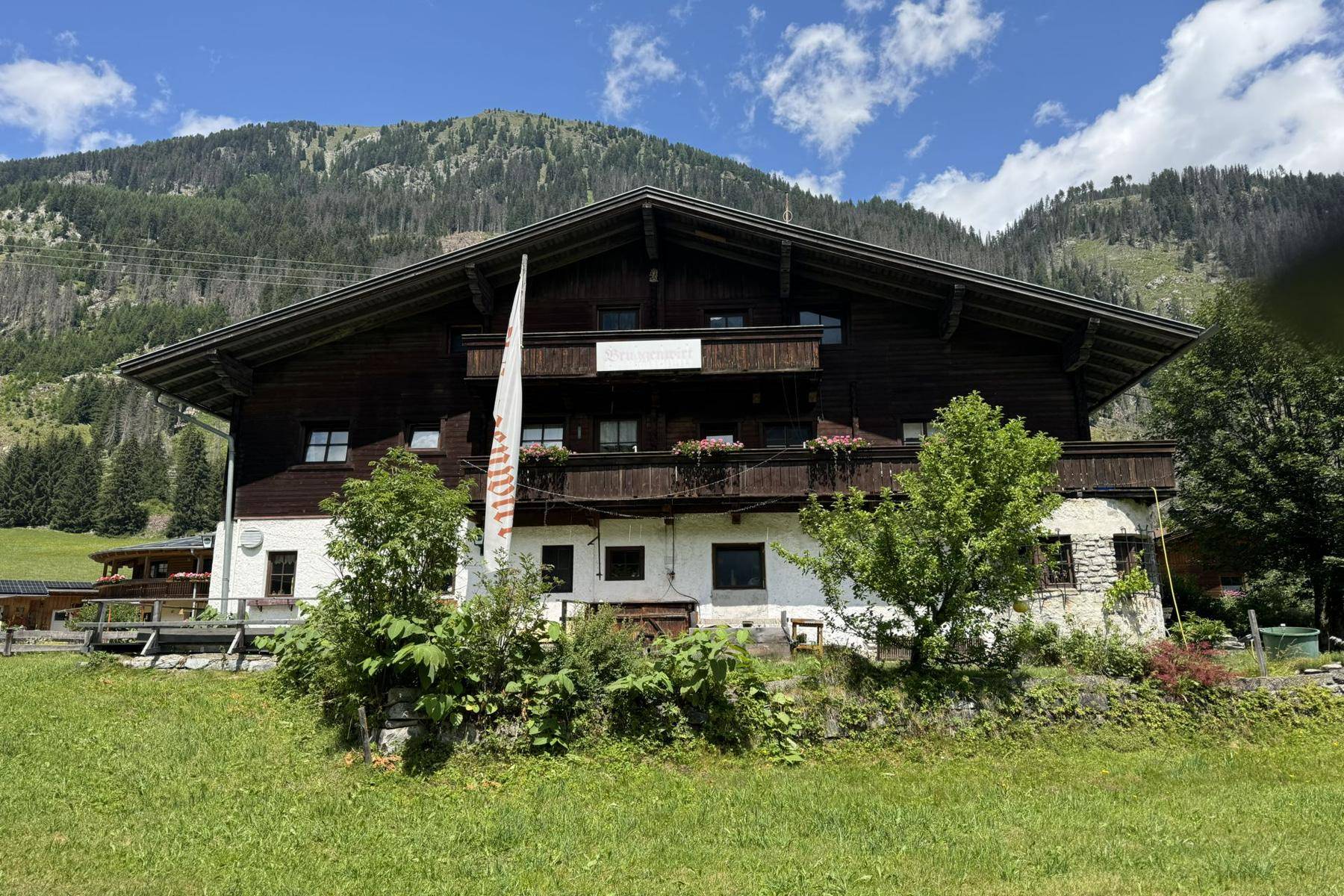 Vorderansicht des traditionellen Holzhauses mit Balkonen und Blumenkästen, umgeben von grünen Wiesen und Bergen.
