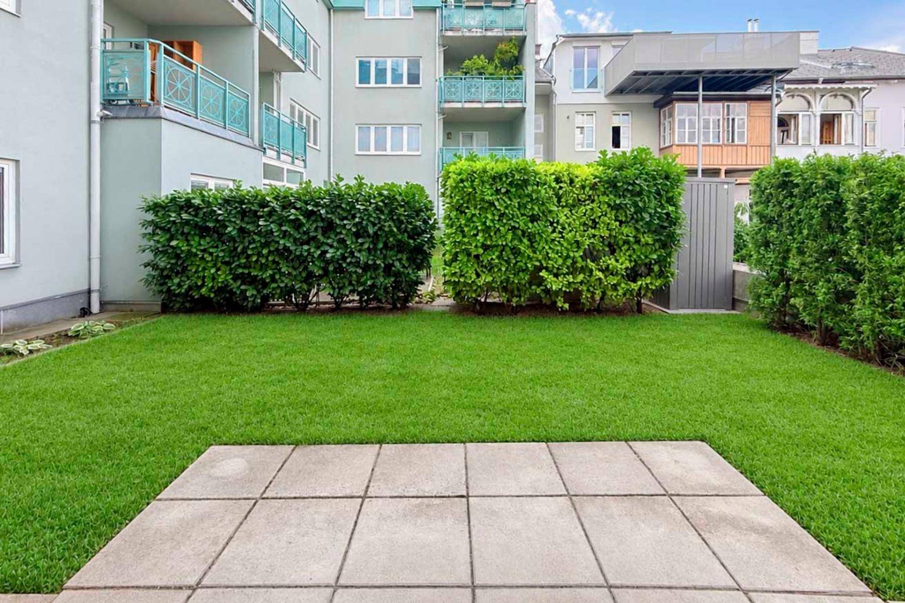 Gepflegter Gartenbereich mit Rasenfläche und gepflasterter Terrasse vor einem modernen Wohngebäude.
