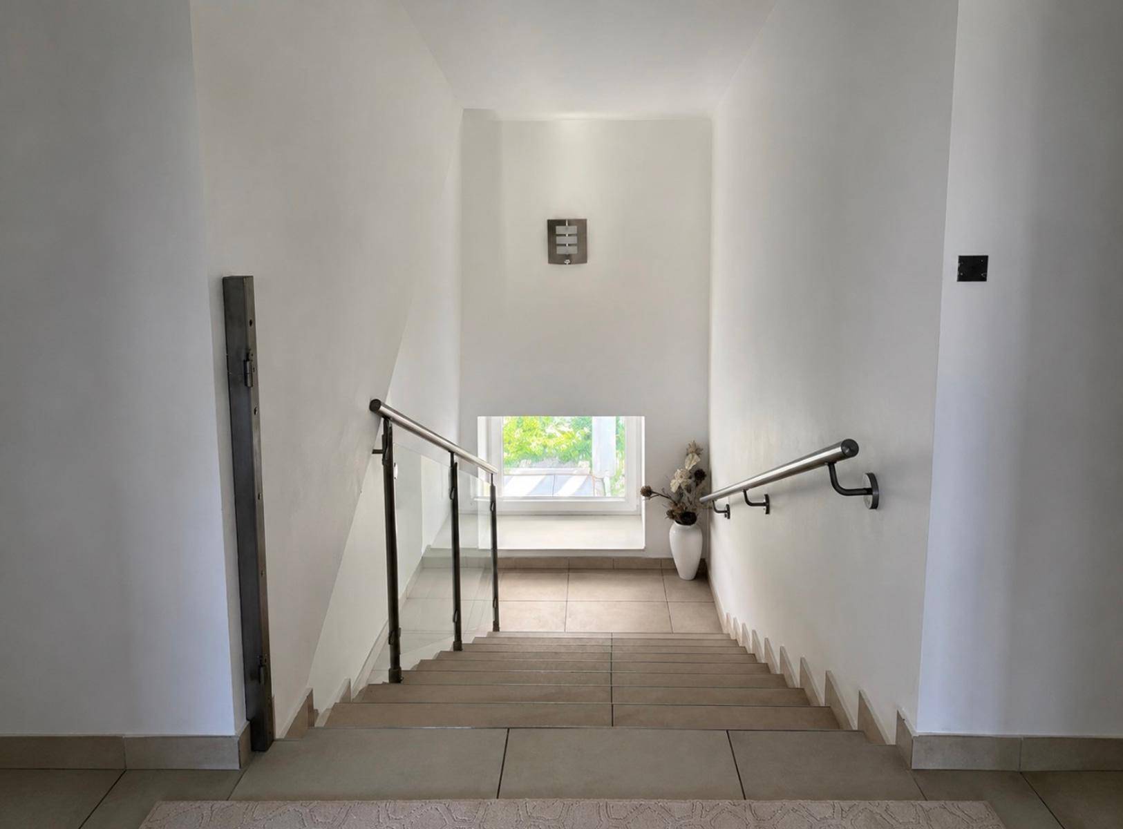 Heller Treppenaufgang mit modernem Geländer und einem Fenster, das Tageslicht hereinlässt.