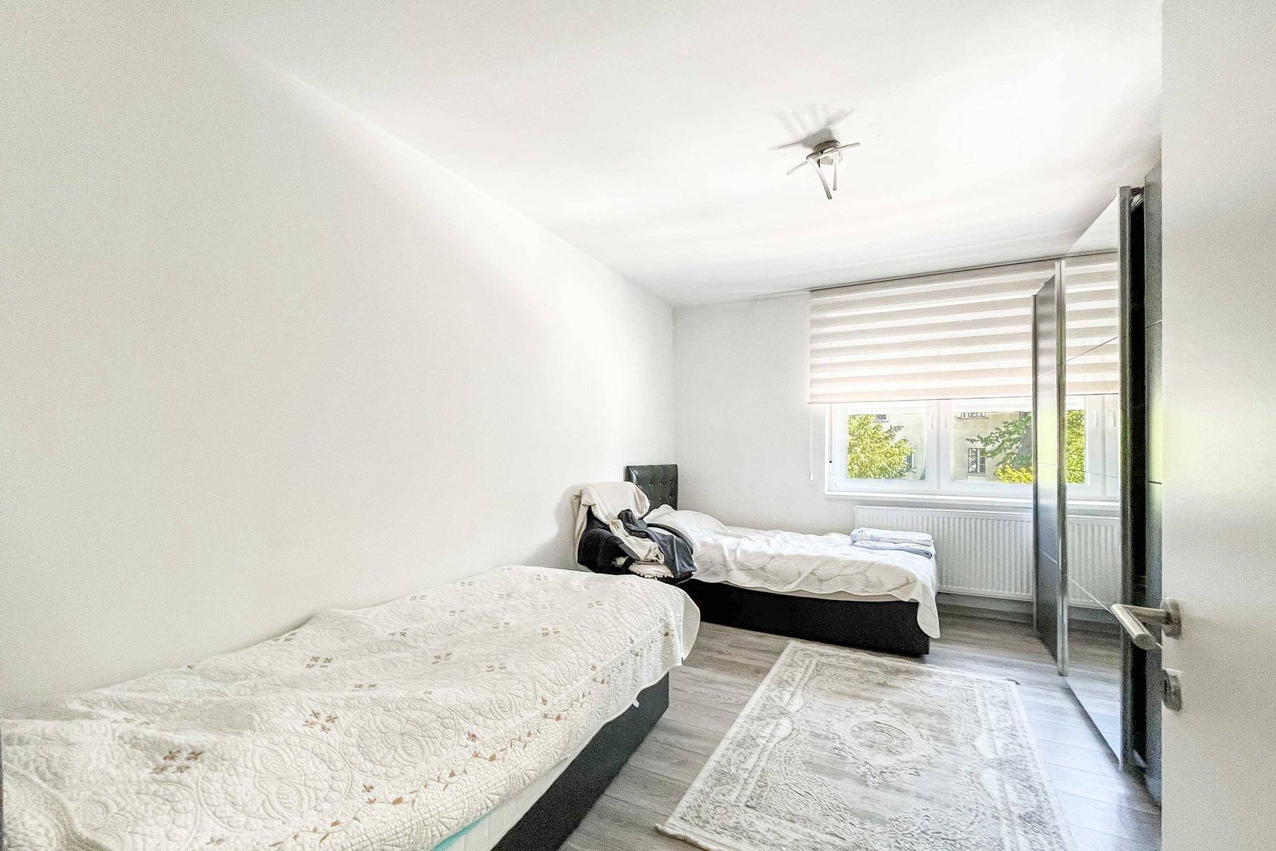 Helles Schlafzimmer mit zwei Betten, modernem Bodenbelag und einem Fenster mit Jalousien.