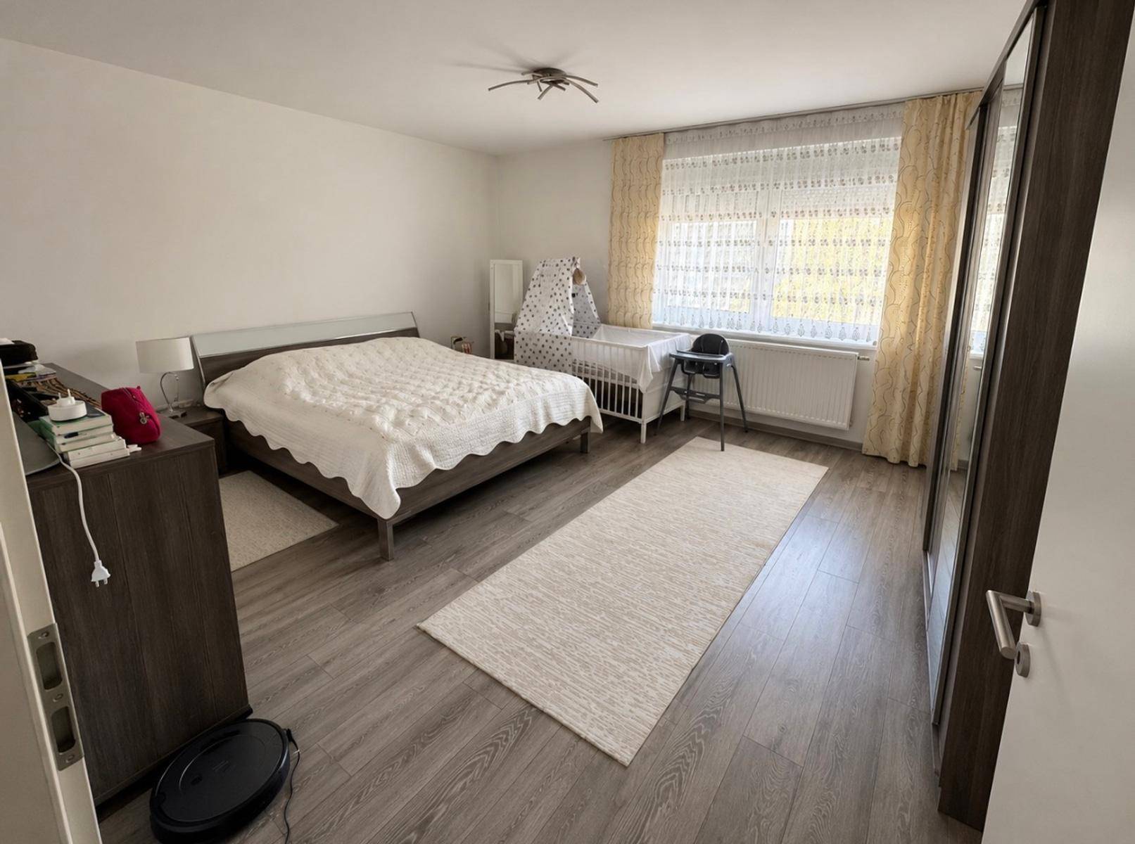 Helles Schlafzimmer mit Doppelbett, Babybett und modernem Bodenbelag, gut beleuchtet.