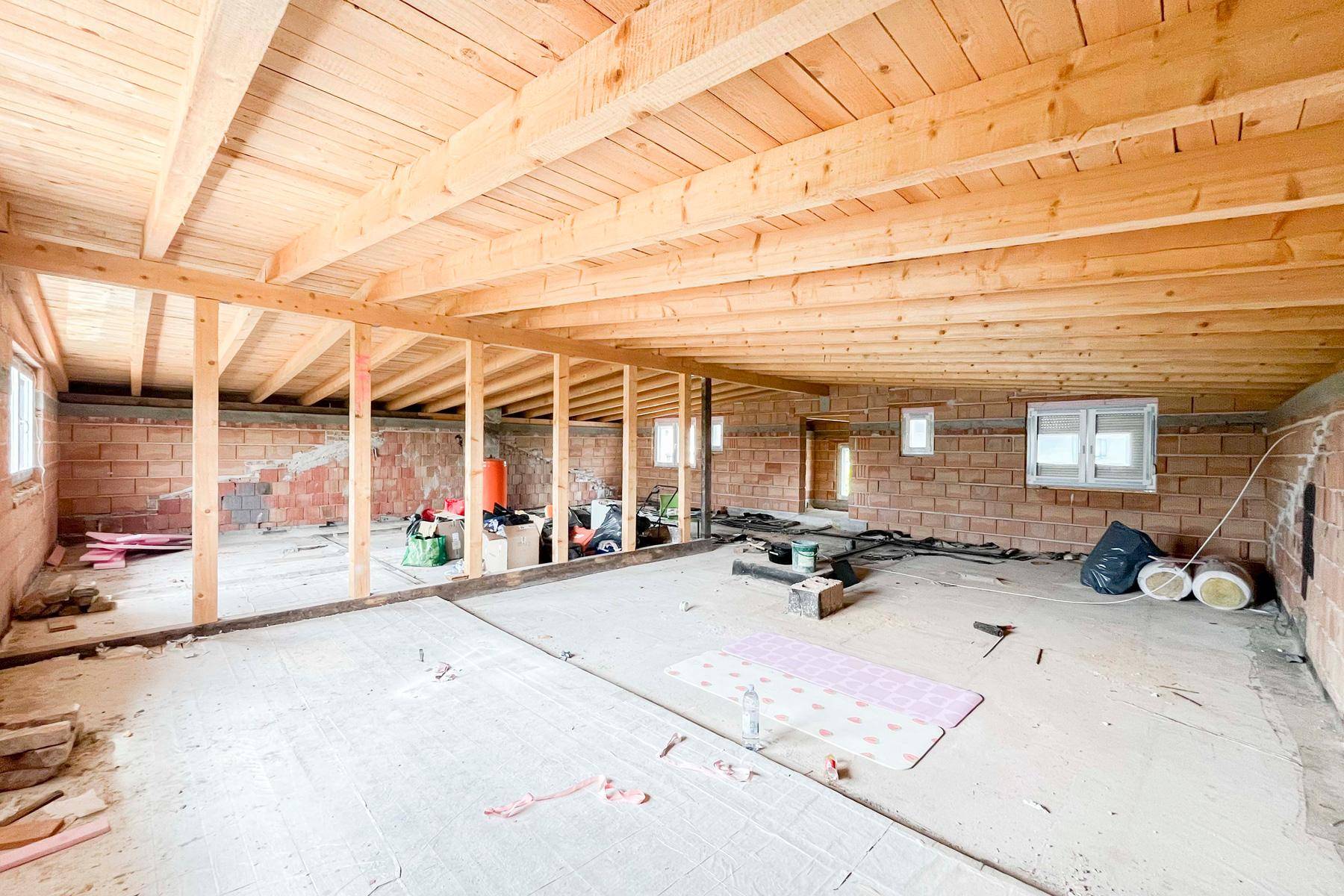 Unfertiger Innenraum im Rohbau mit sichtbaren Holzbalken und Ziegelwänden.