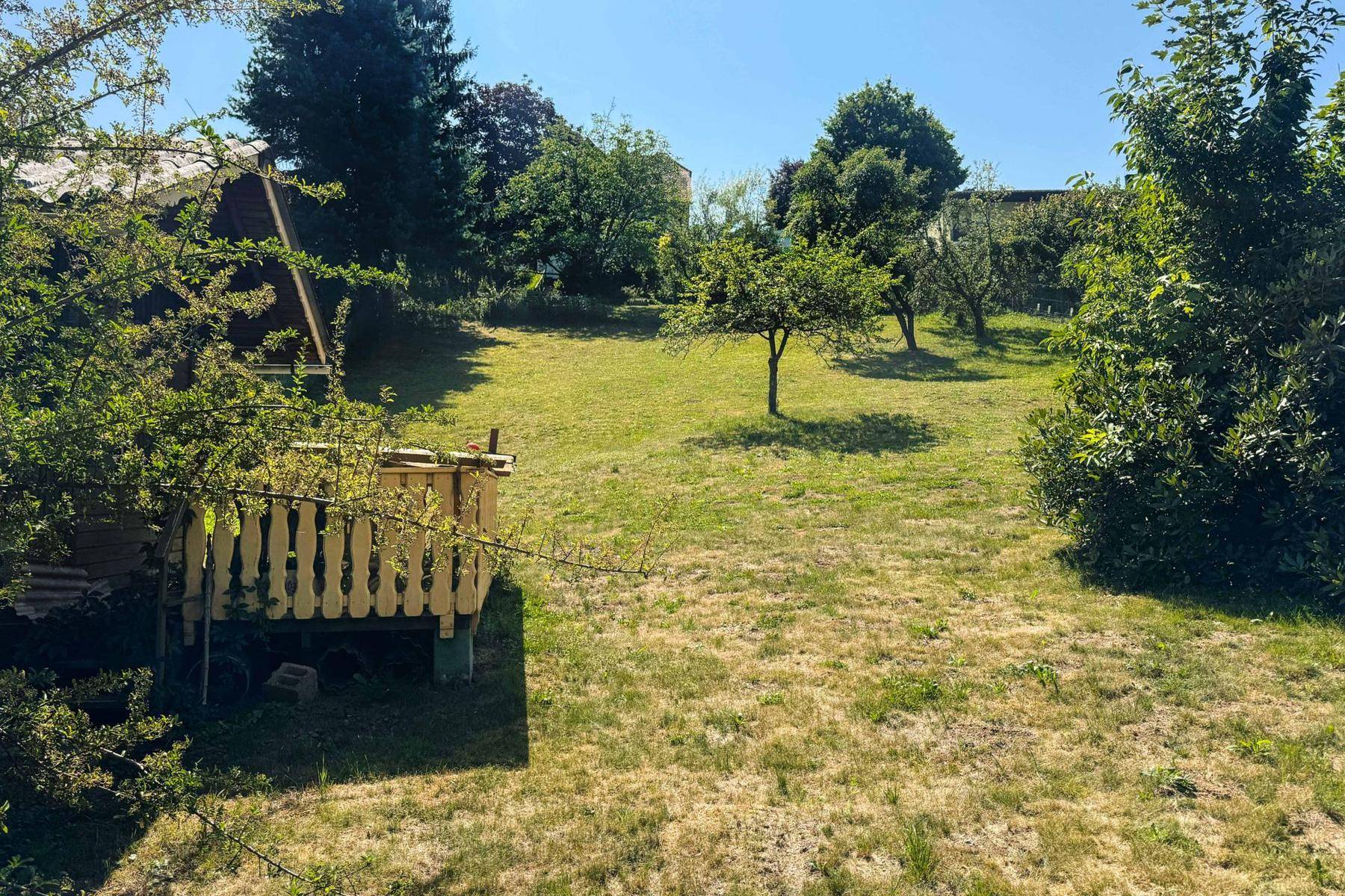 Sonniger Gartenbereich mit Obstbäumen und einem kleinen Holzunterstand im Vordergrund.