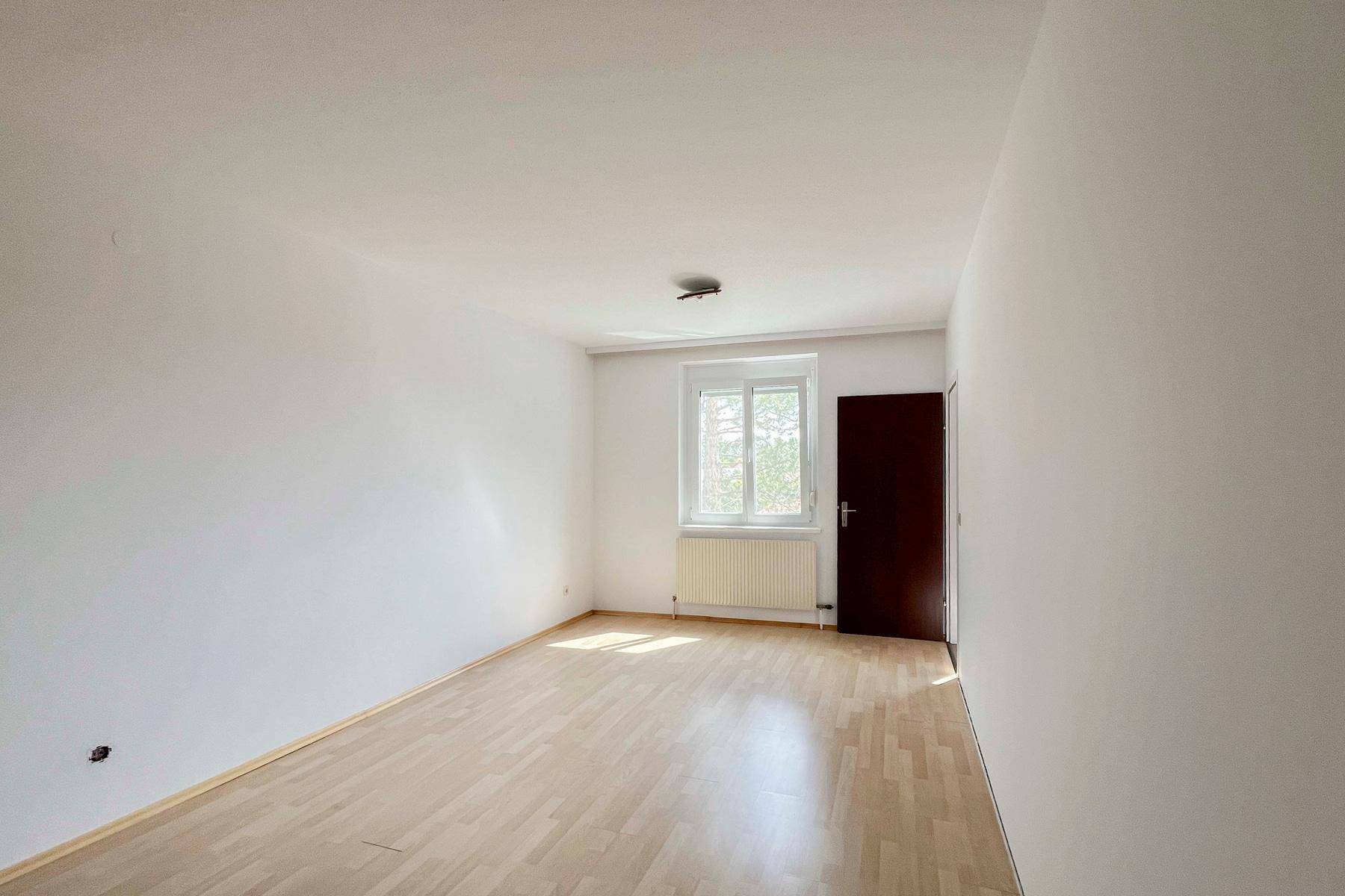 Leerer Raum mit hellem Laminatboden, weißen Wänden und einem Fenster, bereit zur individuellen Gestaltung.