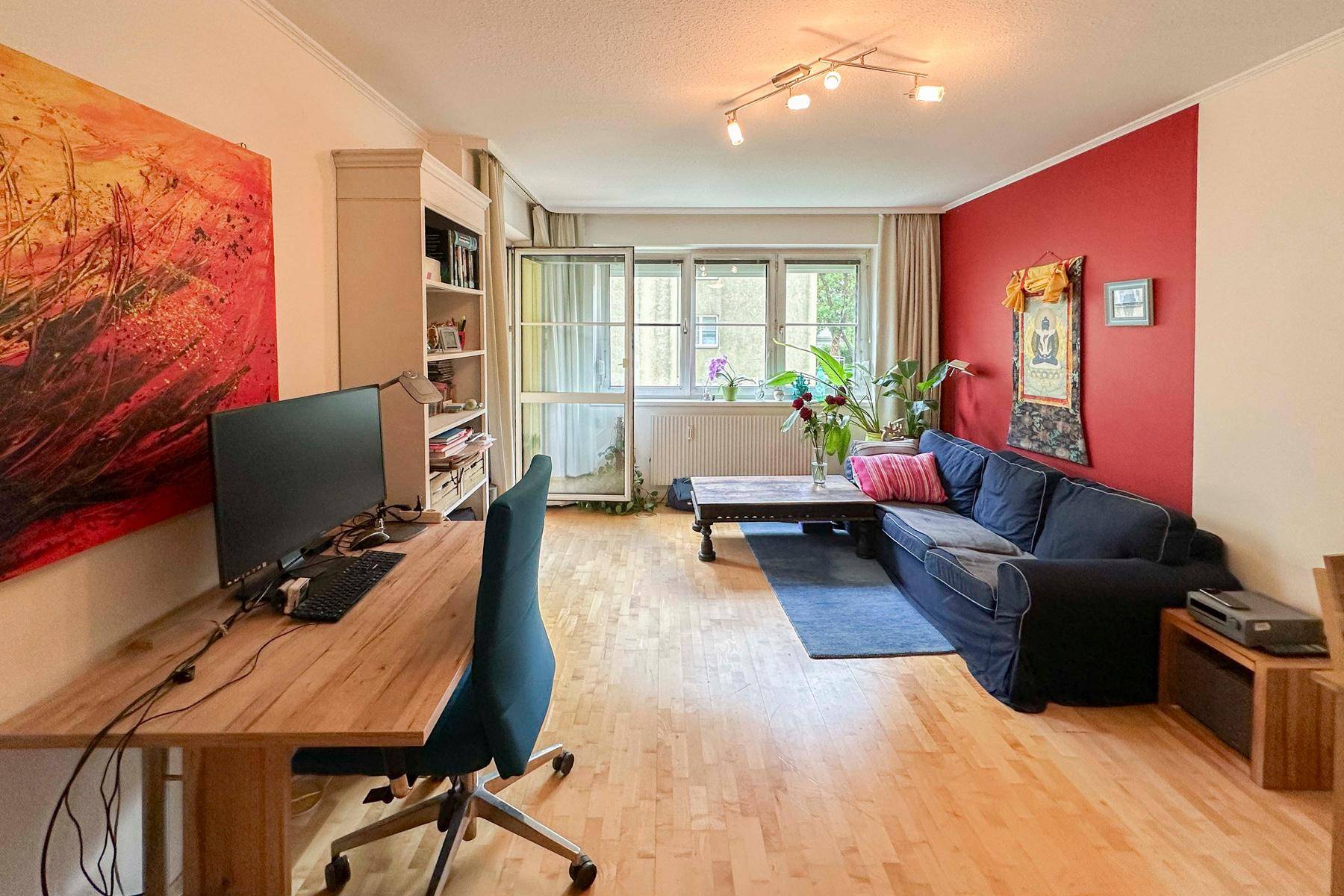 Heller Wohnbereich mit Schreibtisch, Sofa und roter Akzentwand, ideal für Wohnen und Arbeiten.