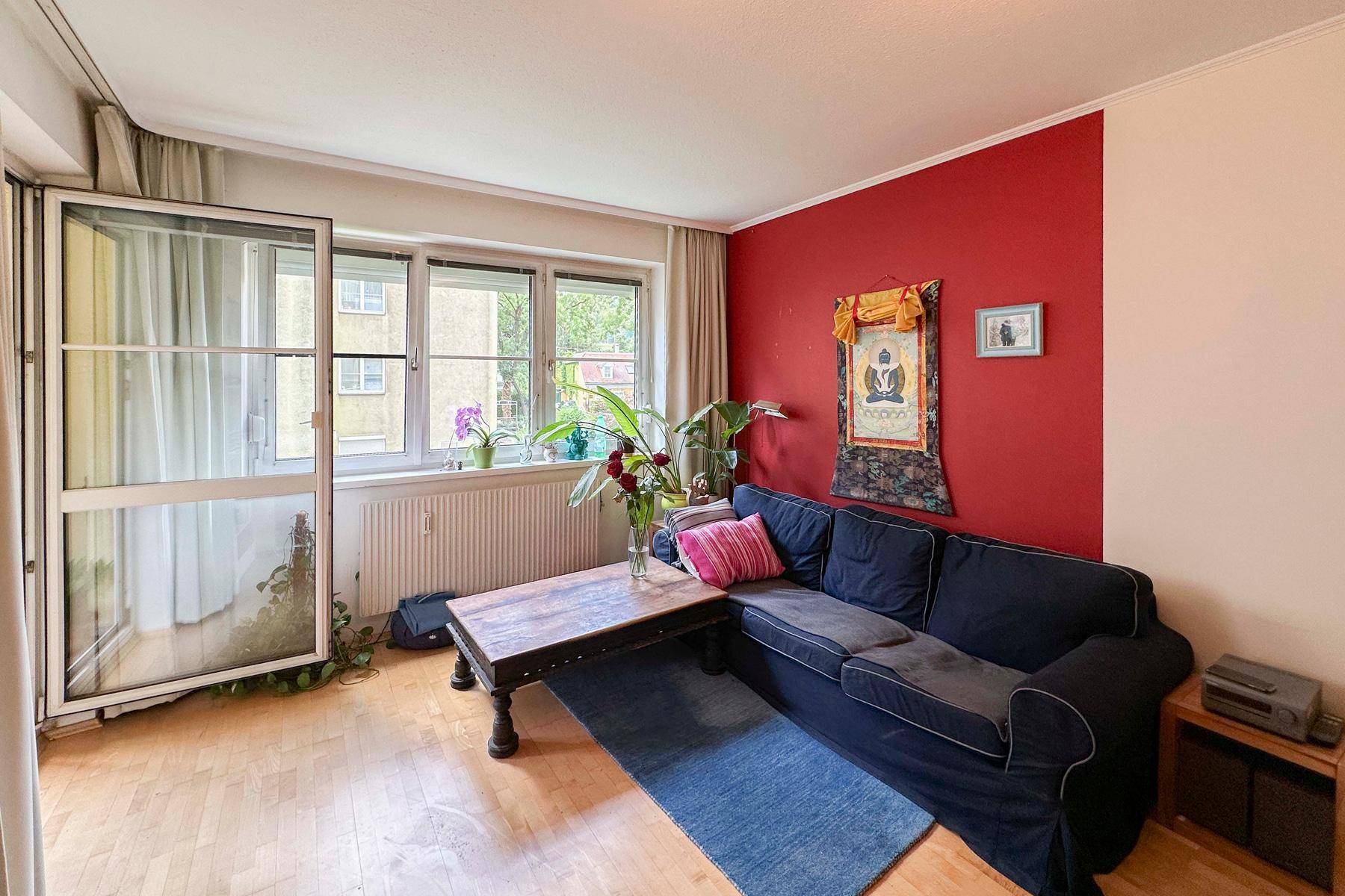 Gemütliches Wohnzimmer mit dunkelblauem Sofa, Holztisch und einer roten Akzentwand, Zugang zum Balkon.