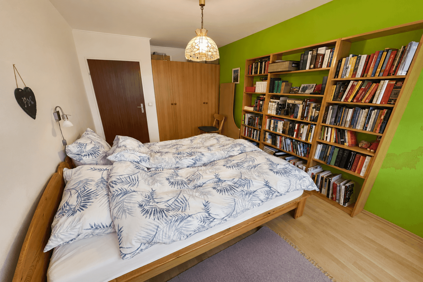 Schlafzimmer mit Holzbett, grüner Akzentwand und einem großen Bücherregal für viel Stauraum.