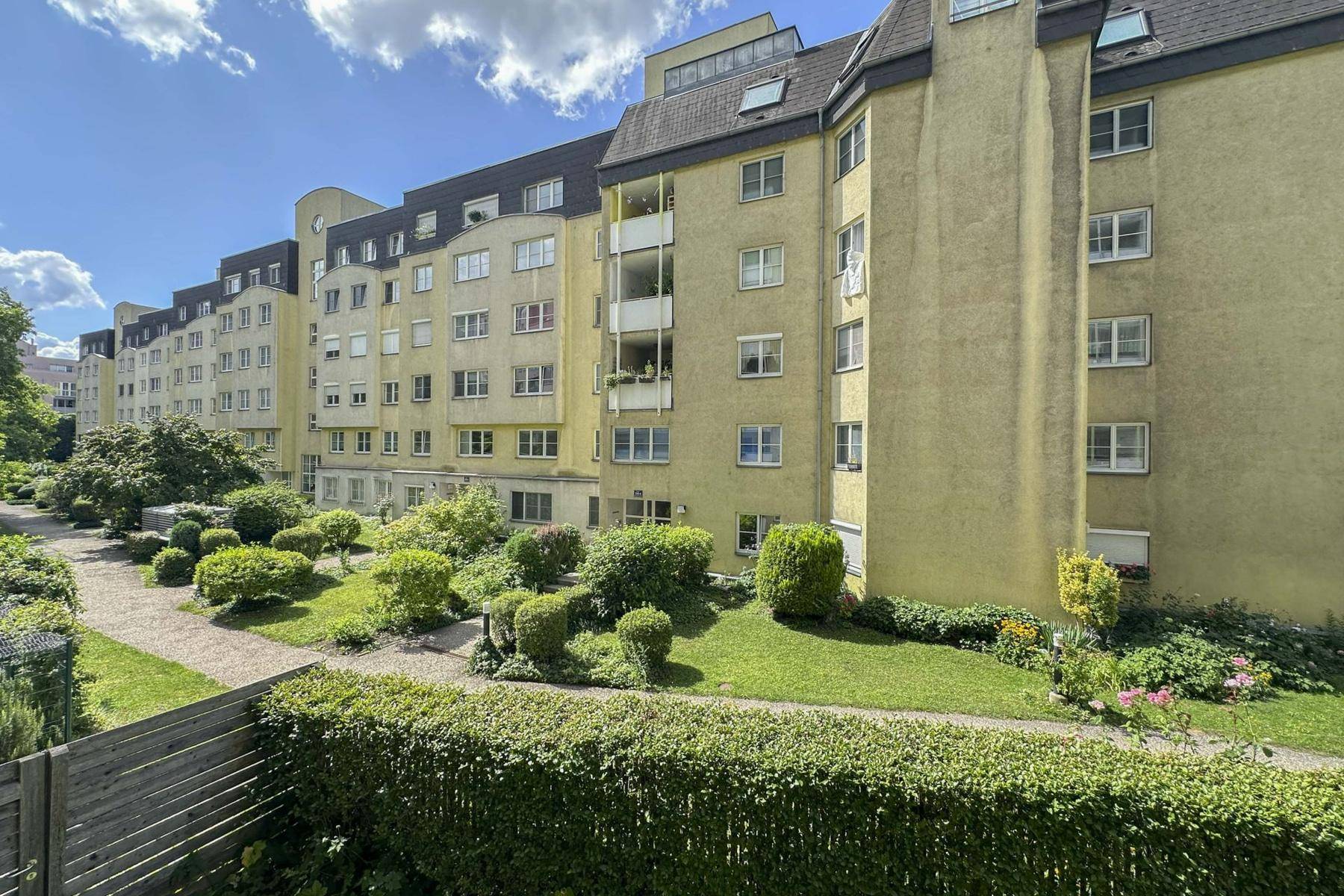 Mehrstöckiges Apartmentgebäude mit gelber Fassade und gepflegtem Gemeinschaftsgarten unter blauem Himmel.