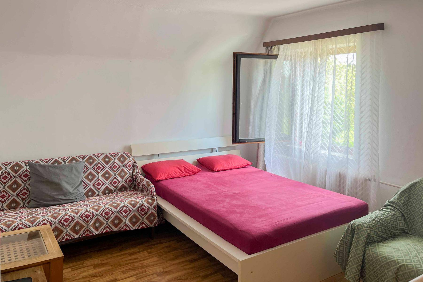 Schlafzimmer mit Holzboden, Sofa und einem Fenster mit Blick ins Grüne.