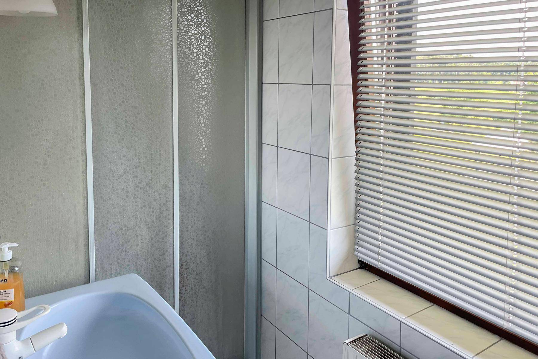 Badezimmer mit Duschkabine, Waschbecken und Fenster mit Jalousien.