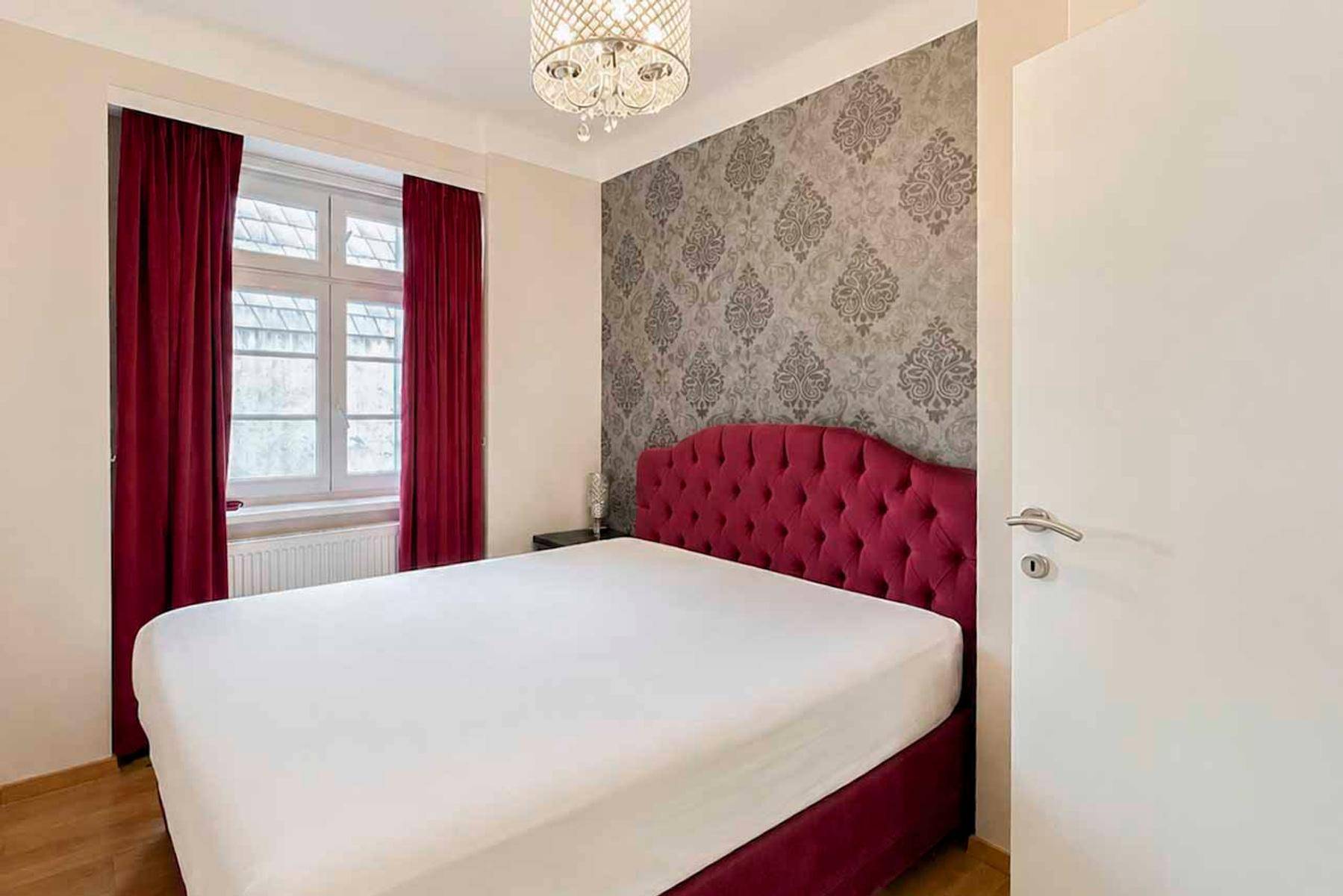 Schlafzimmer mit rotem Polsterbett, gemusterter Tapete und Holzboden.