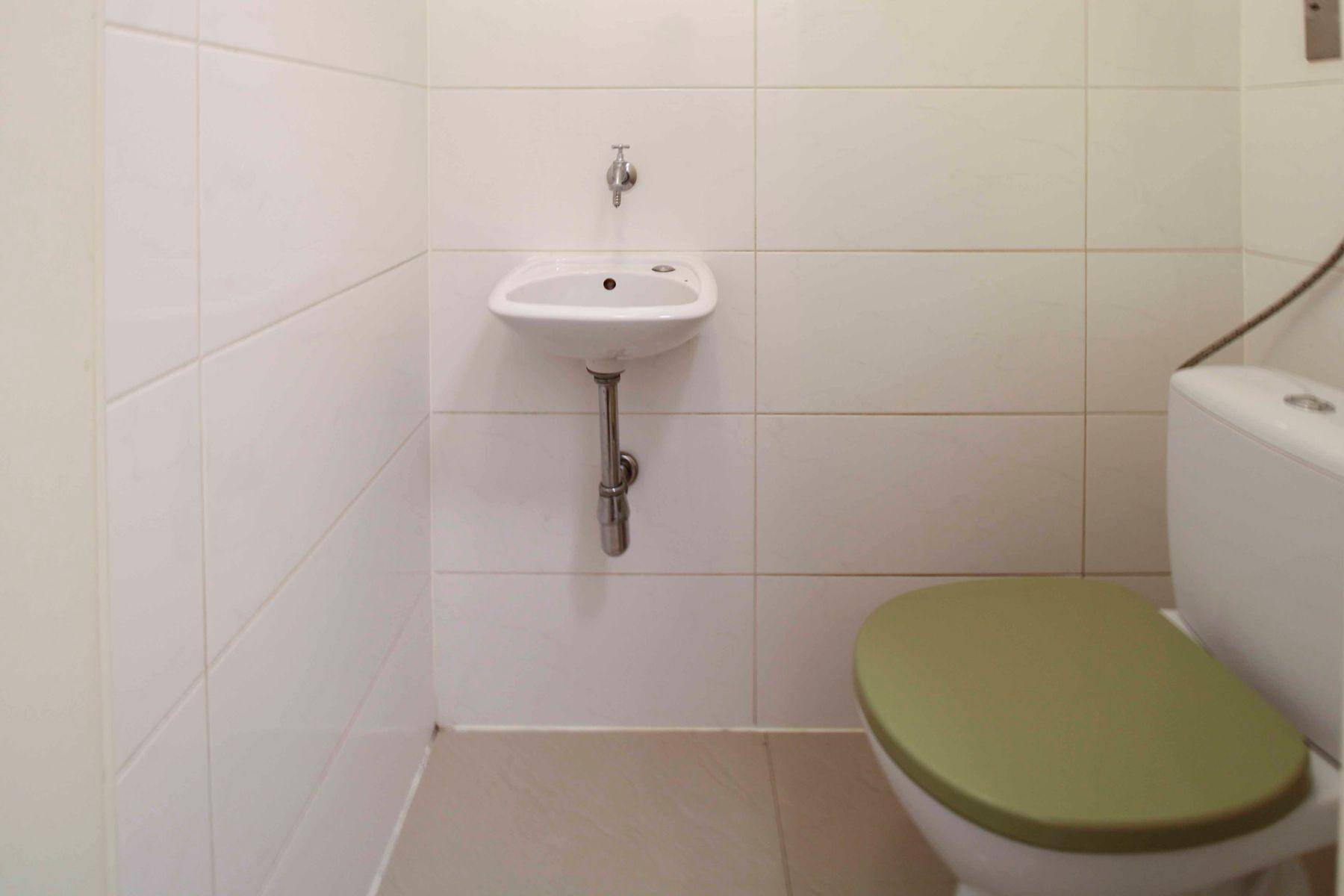 Separates WC mit wandmontiertem Waschbecken und weiß gefliesten Wänden, ausgestattet mit grünem Toilettensitz.