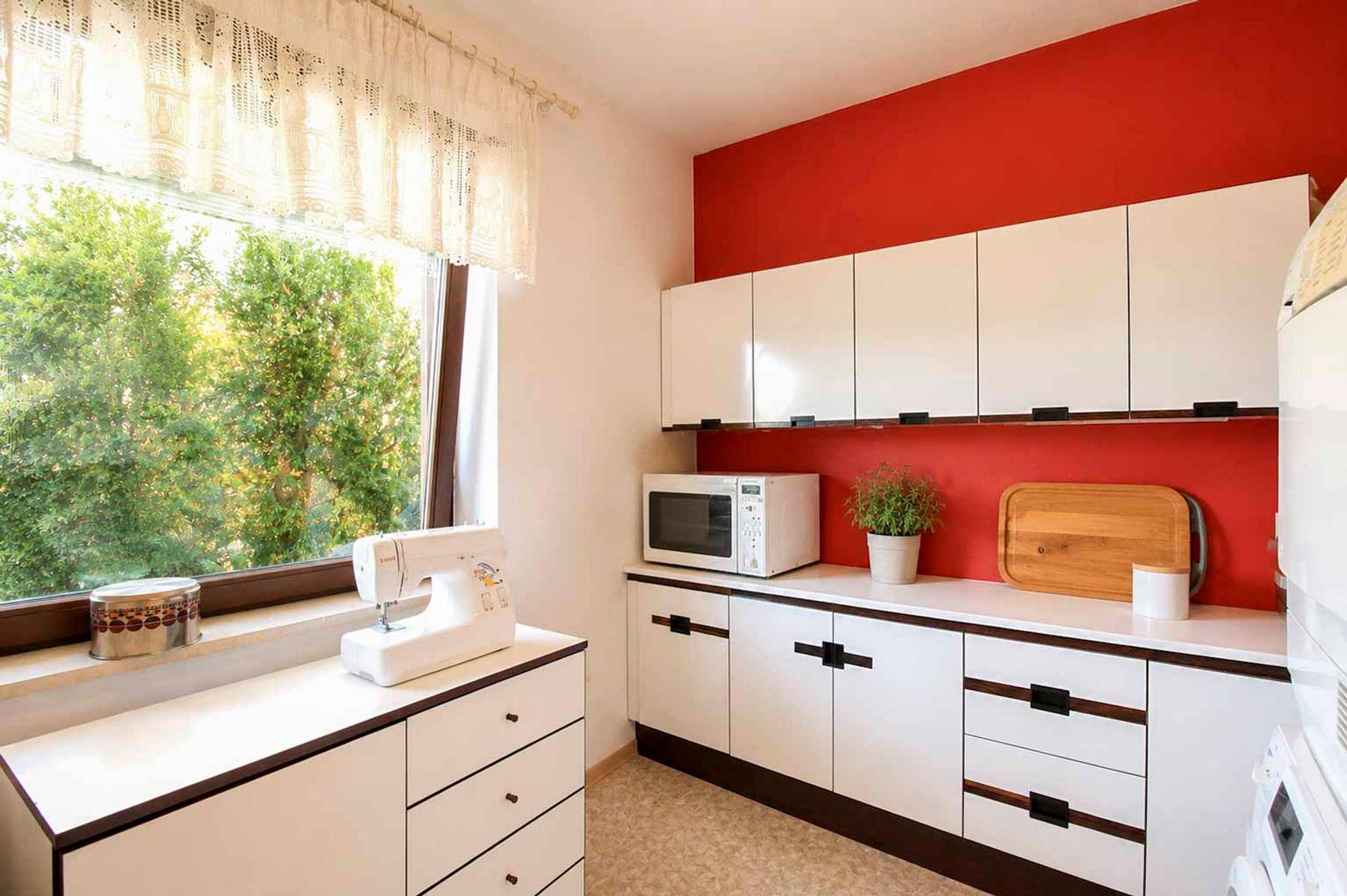 Heller Hauswirtschaftsraum mit weißen Schränken, roter Akzentwand und großem Fenster.