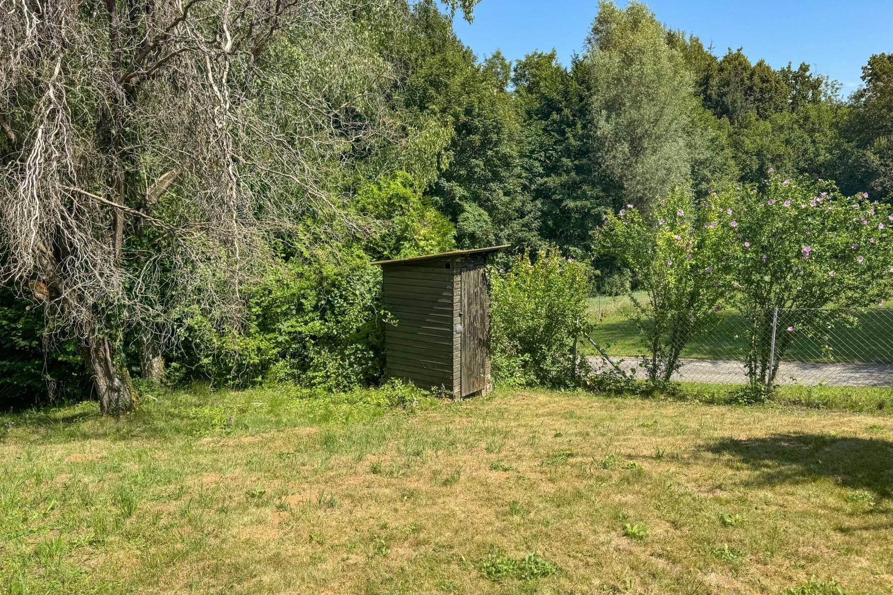 Kleines, rustikales Holzhaus umgeben von üppigem Grün und Bäumen in einem Gartenbereich.