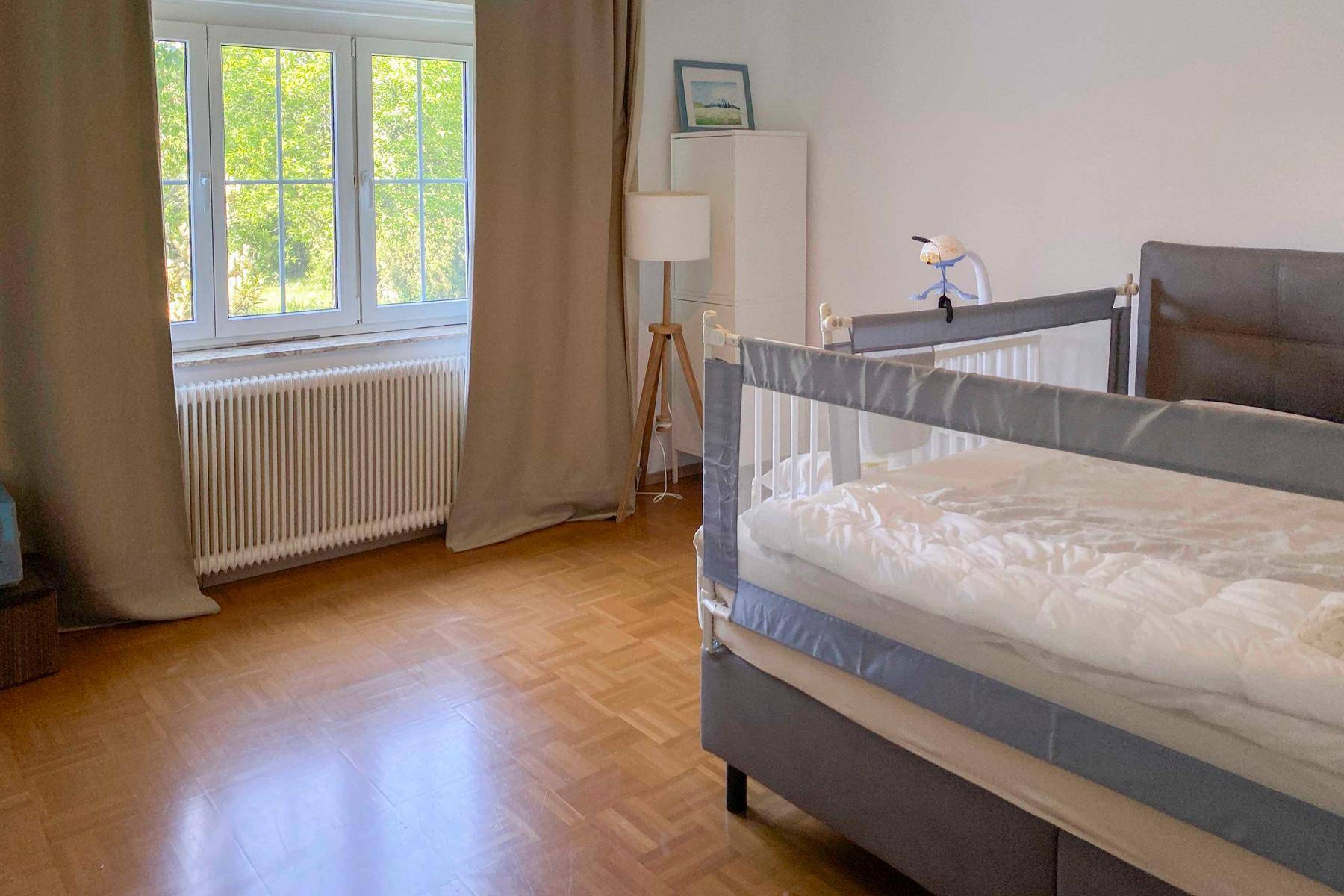 Helles Schlafzimmer mit Parkettboden, großem Fenster und Blick ins Grüne.