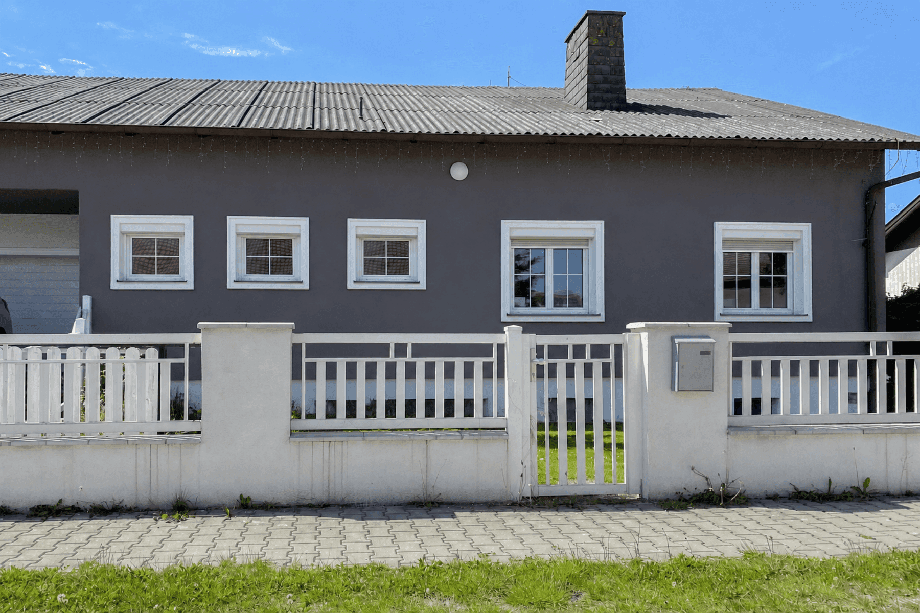 Graue Hausfassade mit weißen Fenstern, einem weißen Zaun und einem Wellblechdach.