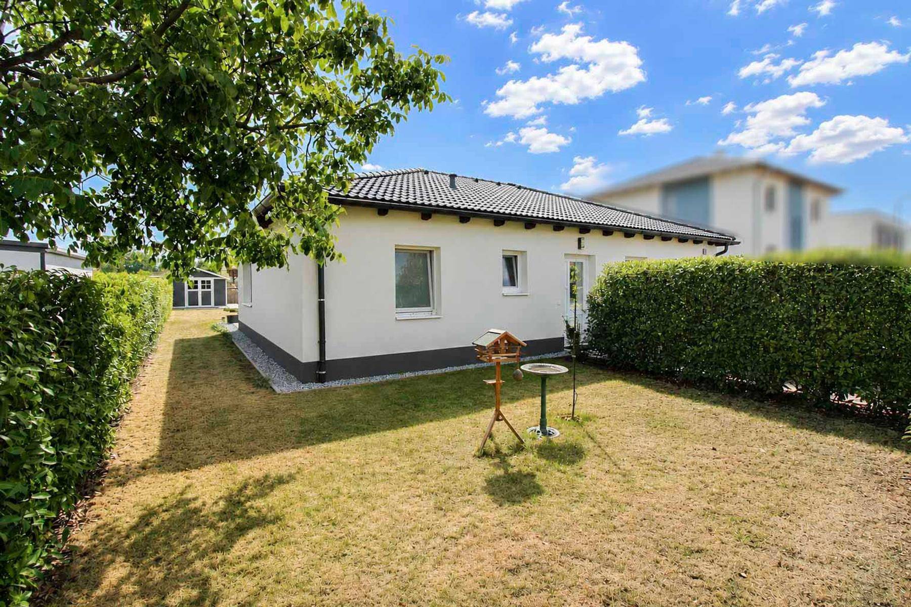 Seitlicher Blick auf das Haus mit hoher Hecke, Rasenfläche und einem kleinen Gartenhaus im Hintergrund.