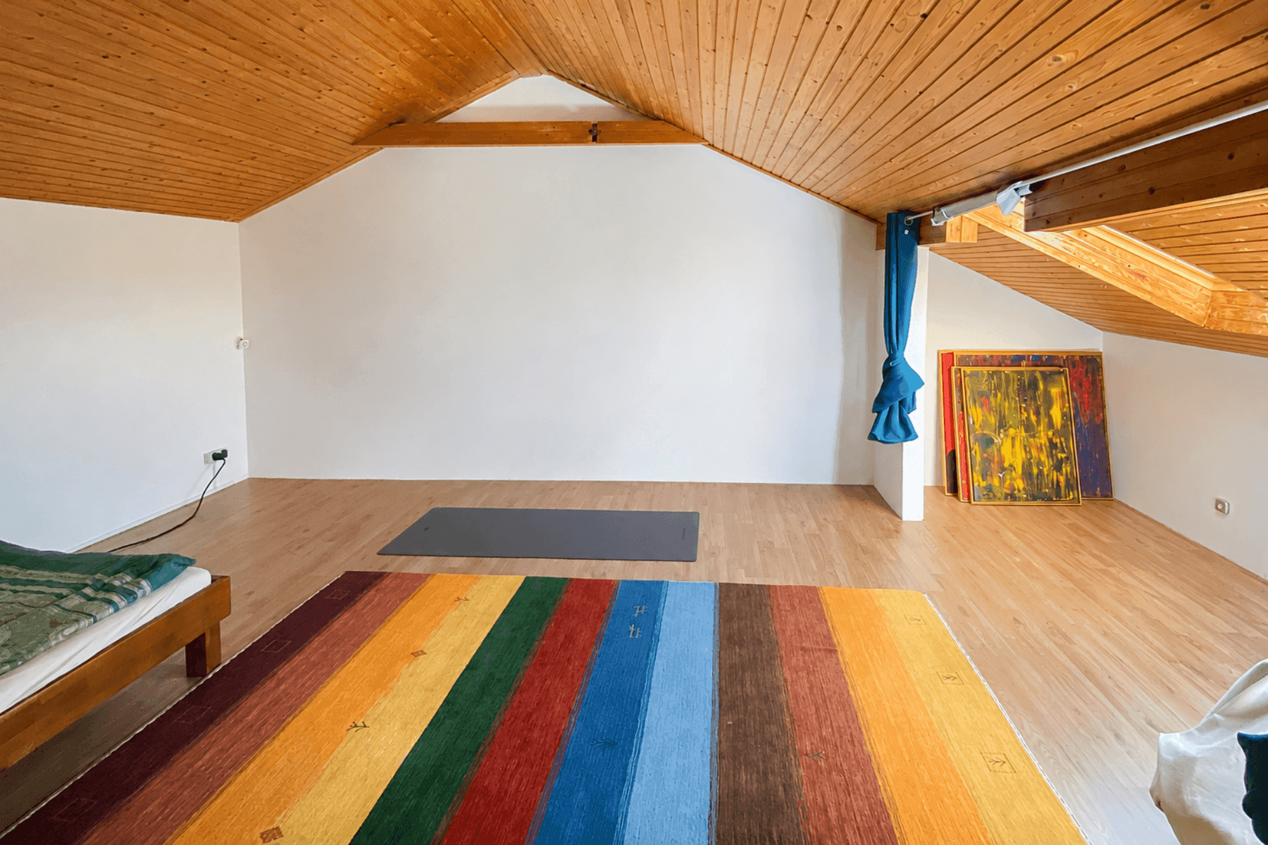 Geräumiges Dachzimmer mit sichtbaren Holzbalken, schrägen Decken und farbenfrohem Teppich.