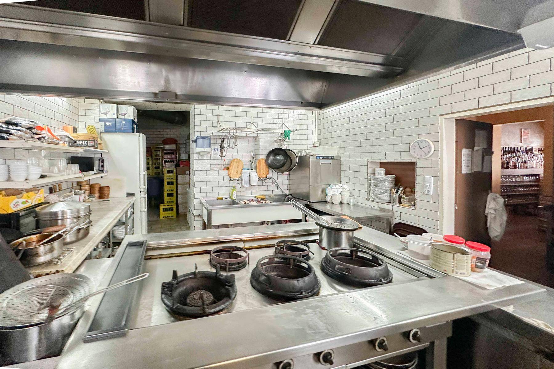 Professionelle Restaurantküche mit Edelstahlgeräten, großen Gasbrennern, weißen Fliesenwänden und einer Dunstabzugshaube.