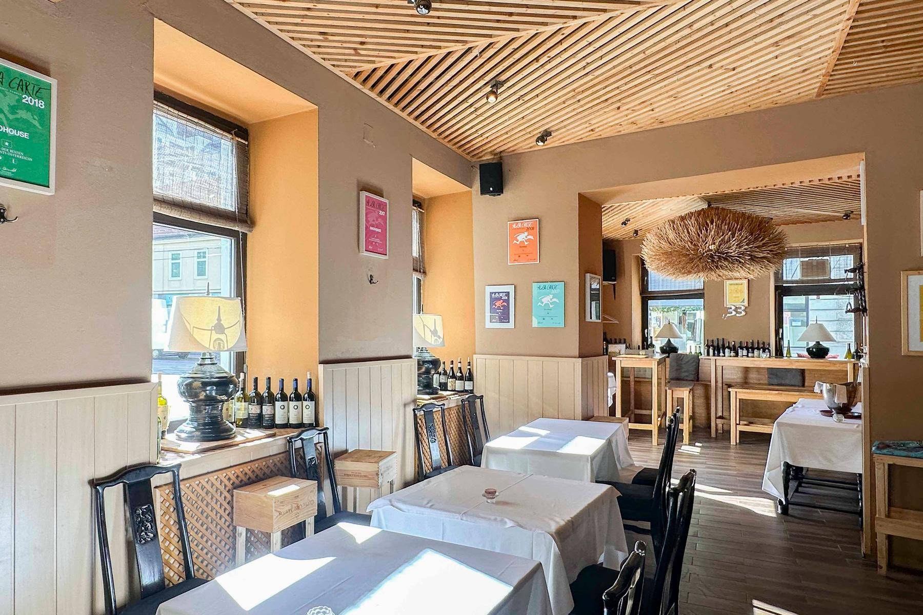 Geräumiger Restaurantbereich mit mehreren Tischen, einer Holzlamellendecke und großen Fenstern, die viel Licht hereinlassen.
