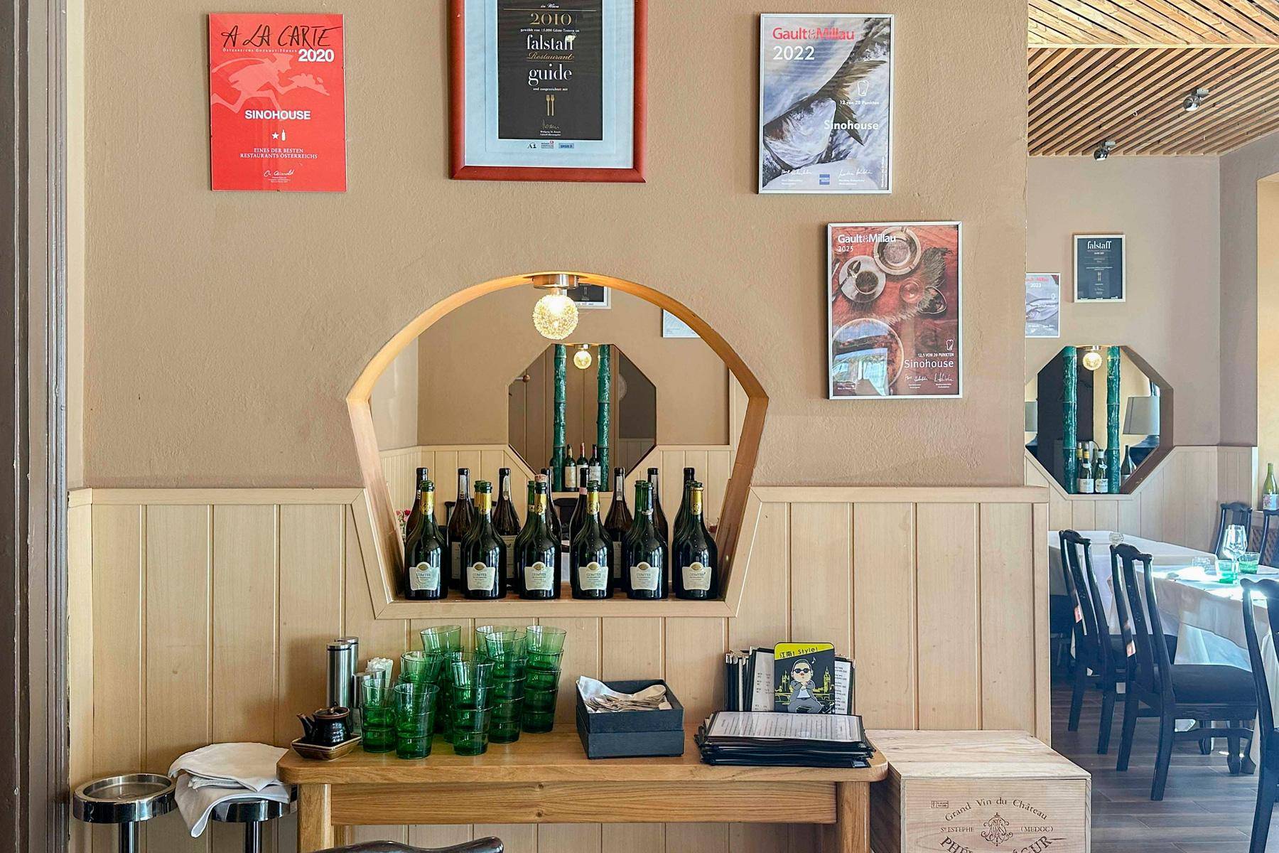 Dekorativer Bereich im Restaurant mit einer Bogennische für Weinflaschen, grünen Gläsern und gerahmten Bildern an der Wand.