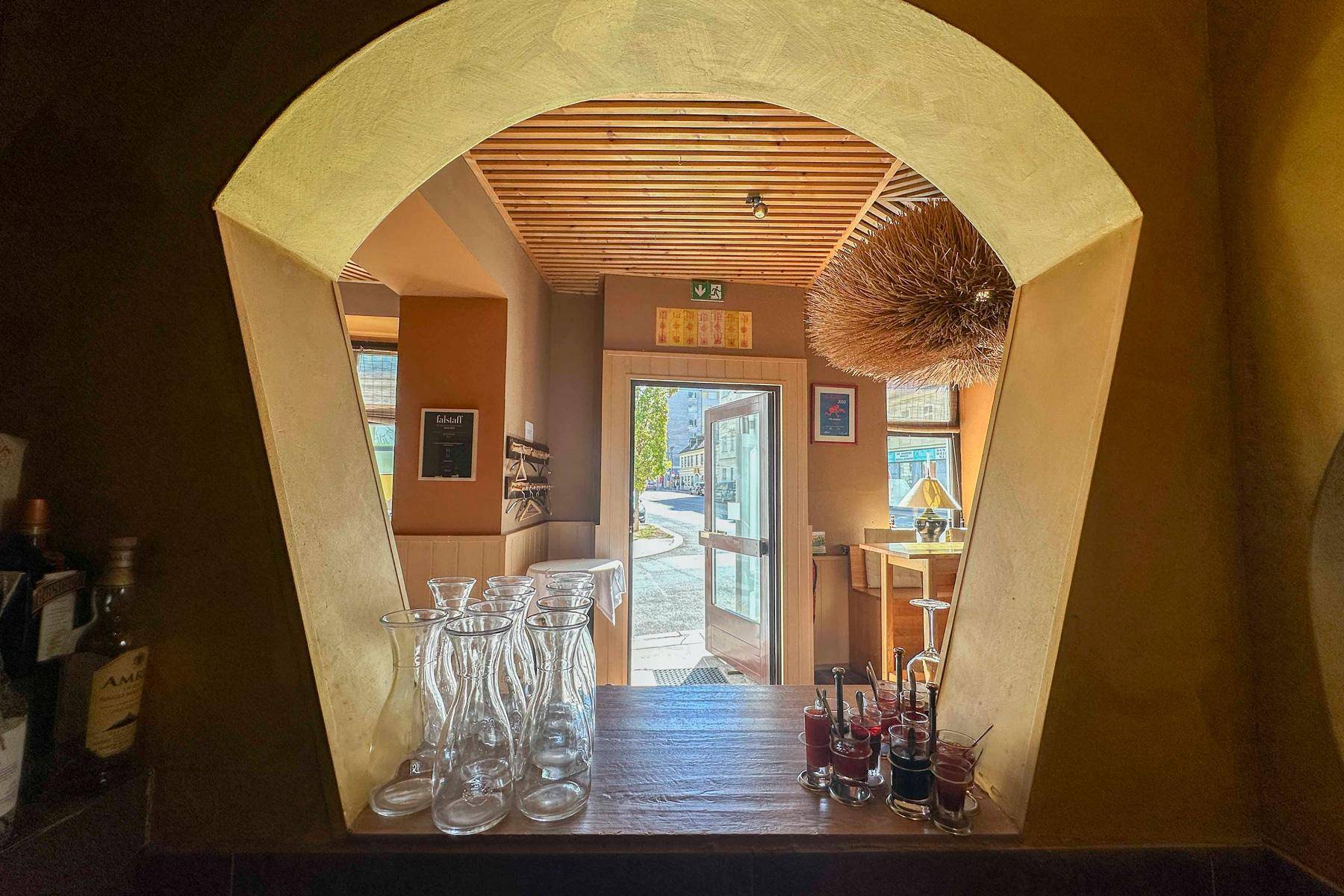 Blick durch eine Bogenöffnung vom Servicebereich in den Hauptraum des Restaurants mit sichtbarem Eingang.