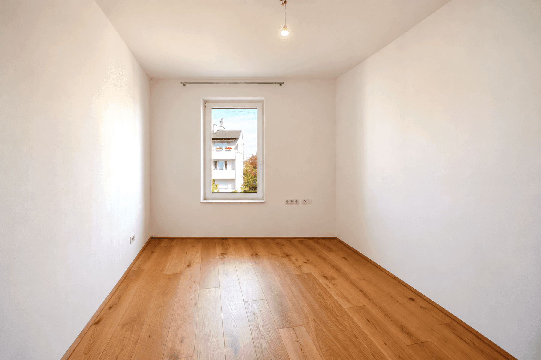 Leerer Raum mit hellem Holzboden und einem Fenster, das den Blick nach draußen freigibt.