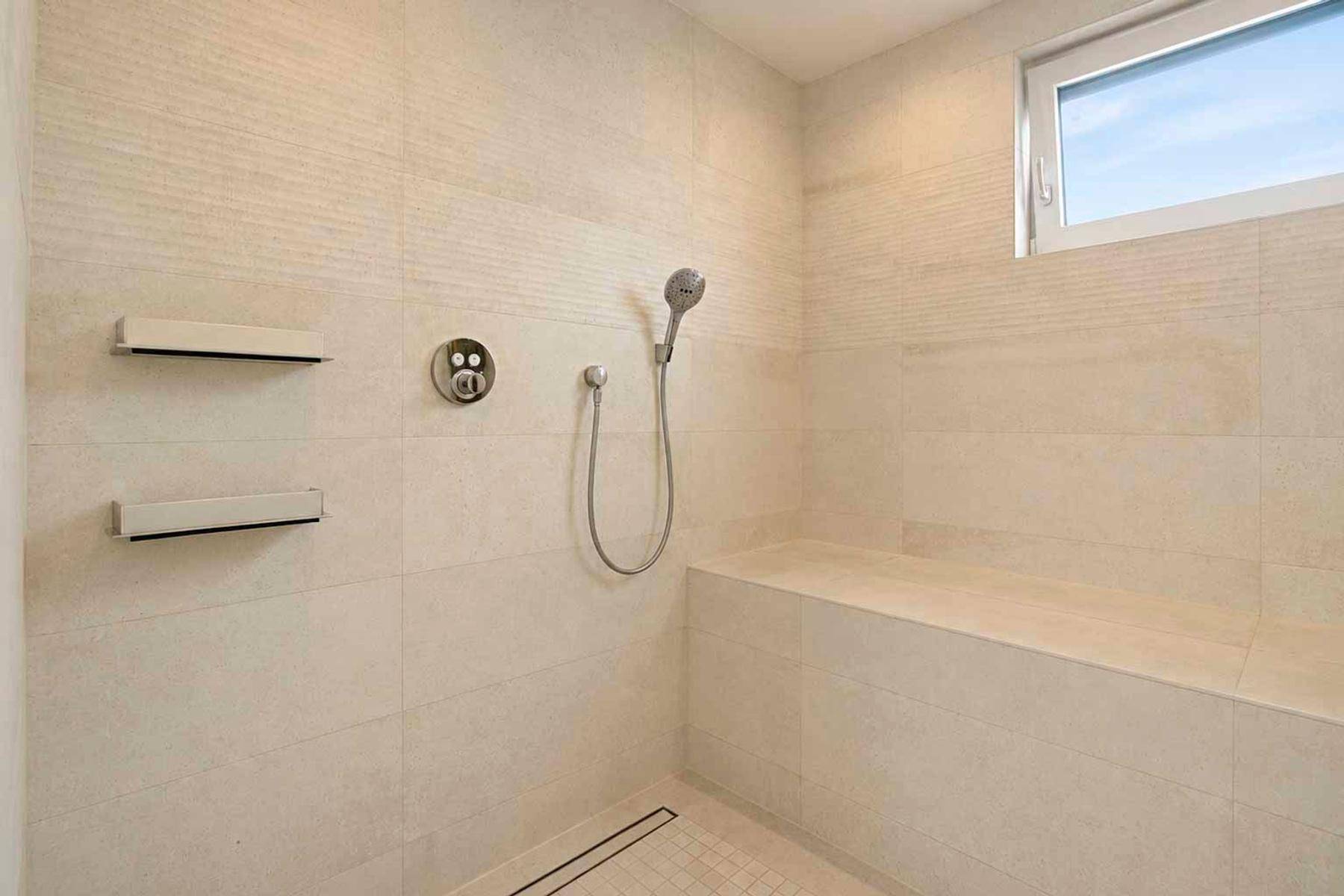 Große begehbare Dusche mit hellen Fliesen, integrierter Sitzbank und Fenster für Tageslicht.