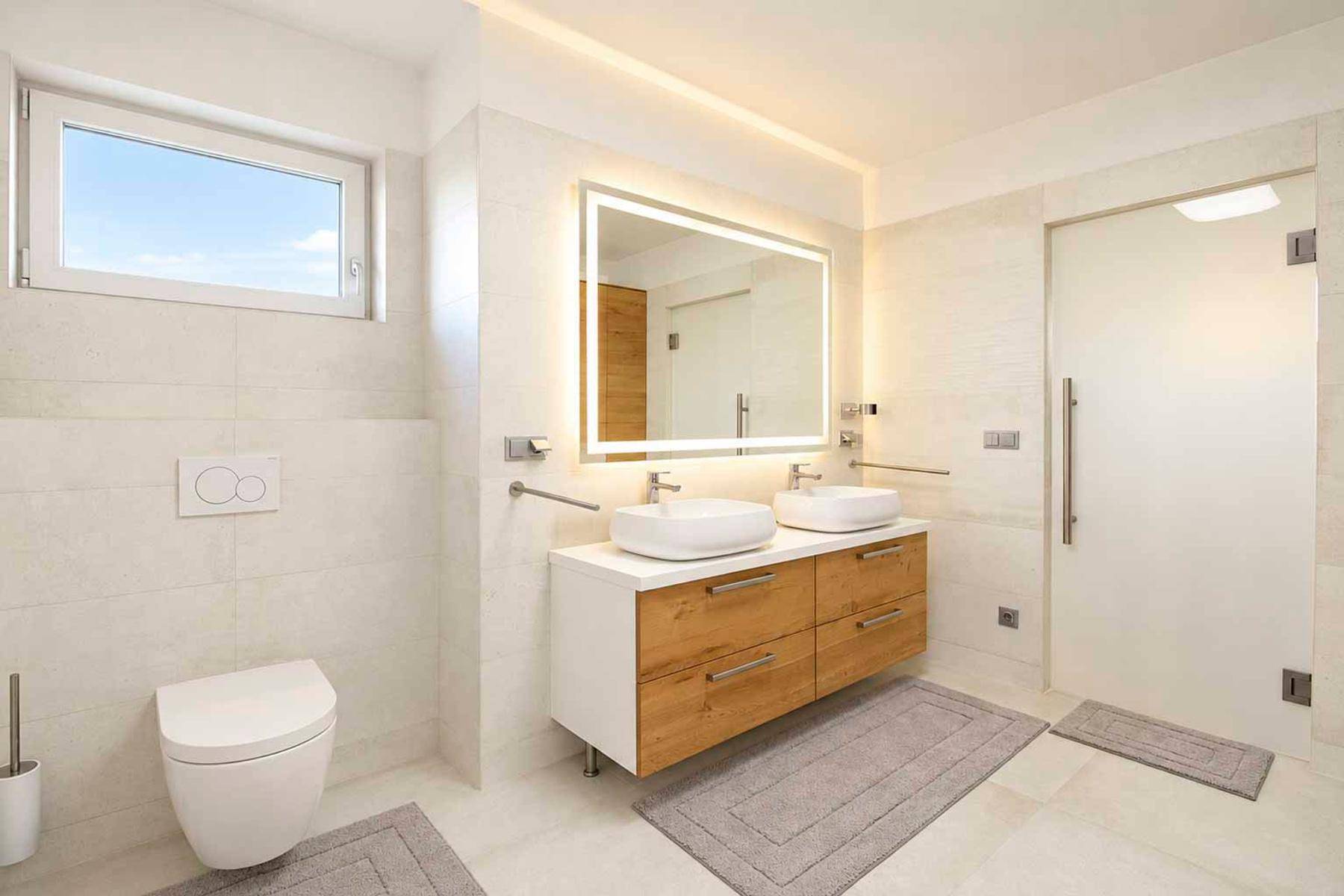 Geräumiges modernes Badezimmer mit Doppelwaschbecken, beleuchtetem Spiegel und hellen Fliesen.
