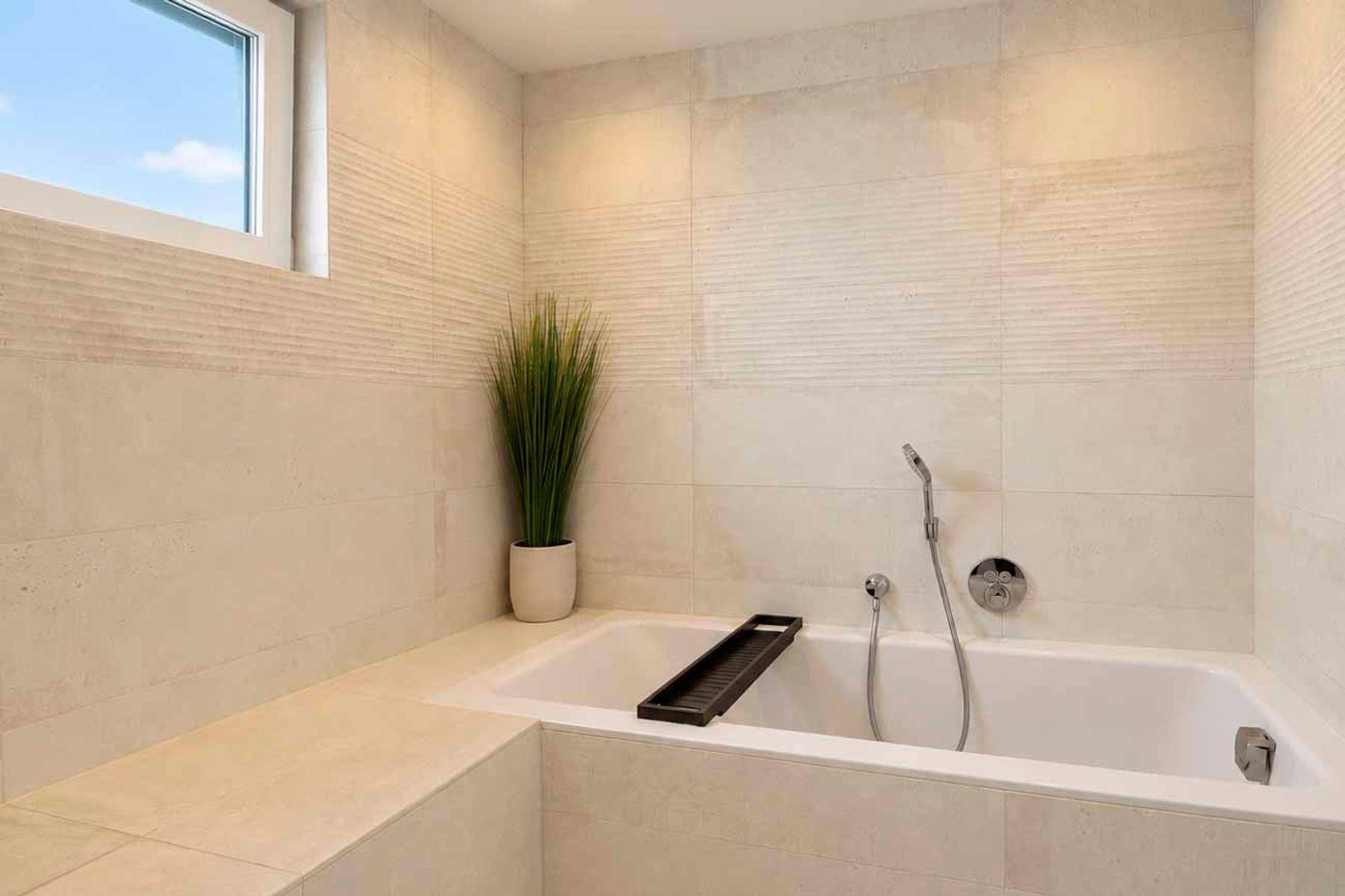 Elegante Badewanne mit hellen Fliesen, integrierter Ablagefläche und Fenster für natürliches Licht.