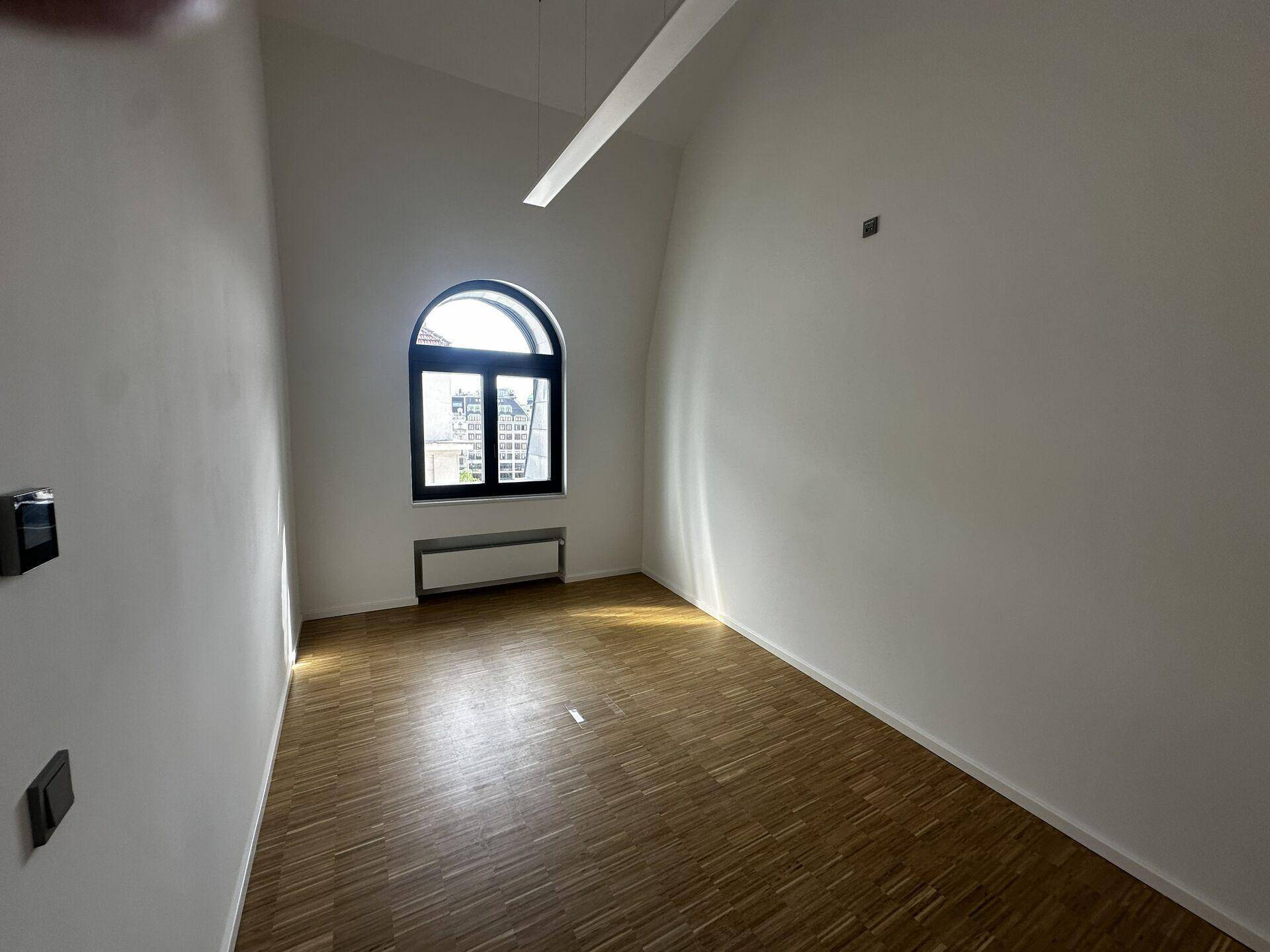 Heller Büroraum mit Holzboden, einer modernen Deckenleuchte und einem eleganten Rundbogenfenster.